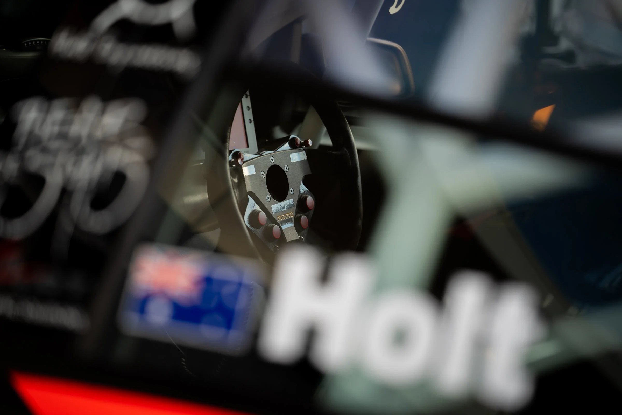 Close-up view through the window of a race car interior, showing a momo steering wheel and the name ‘Holt’ on the glass, with the roll cage visible behind