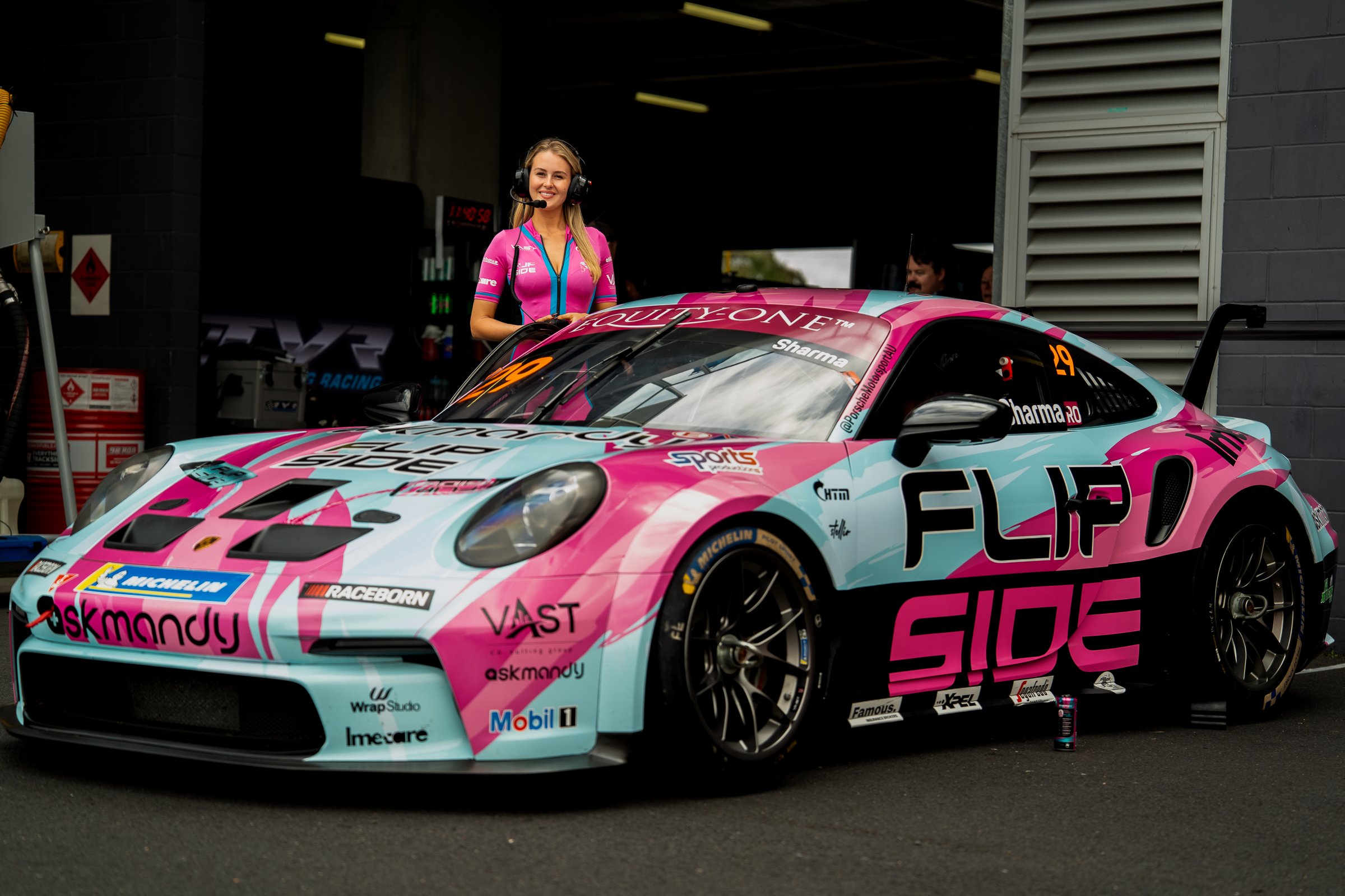 Promotional model in a pink and blue bodysuit wearing a headset smiling beside a pink and blue Porsche race car in front of a garage