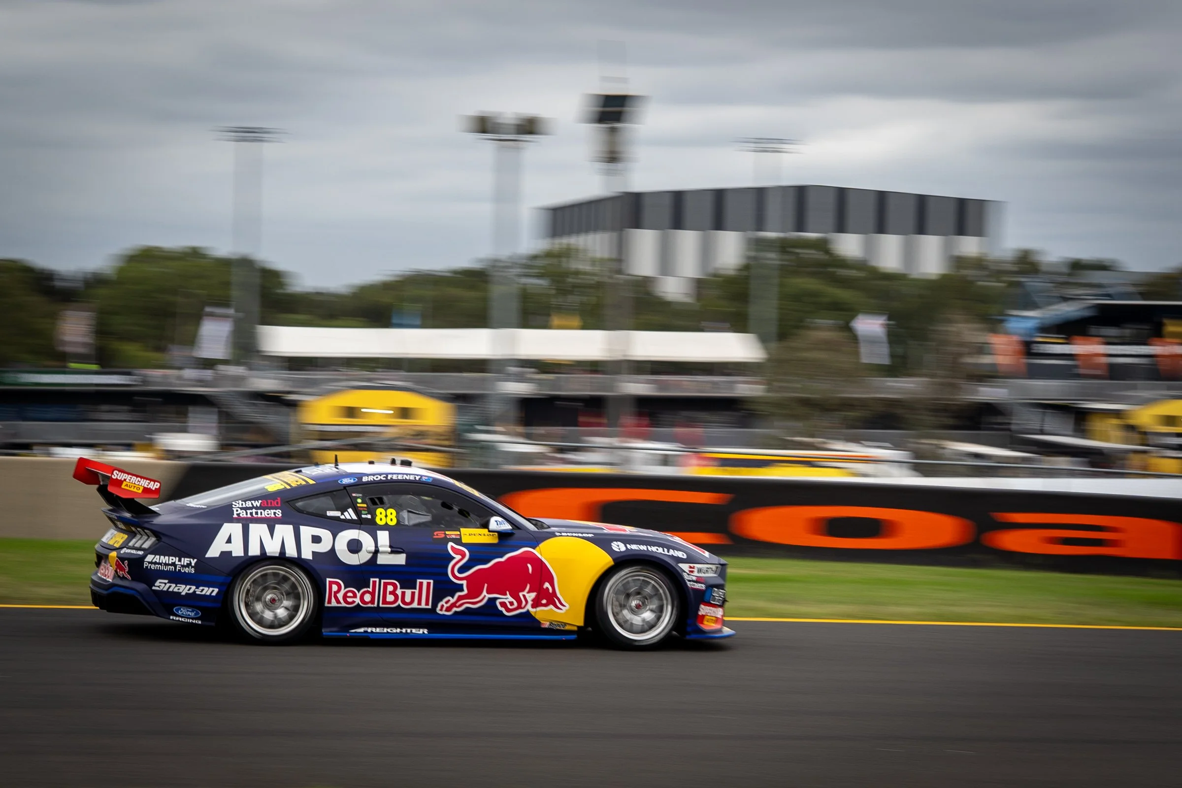 Red Bull Ampol–branded race car speeding along the racetrack with blurred grandstands and an overcast sky in the background