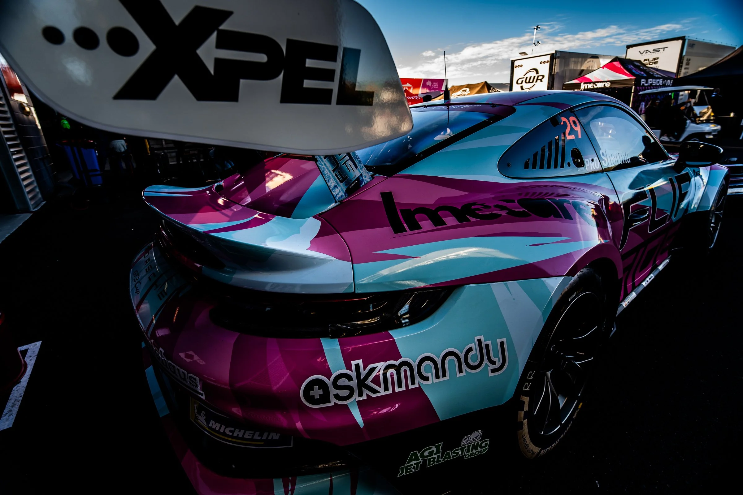 Porsche race car with pink, light blue and white livery parked in a paddock area with tents and trailers in the background