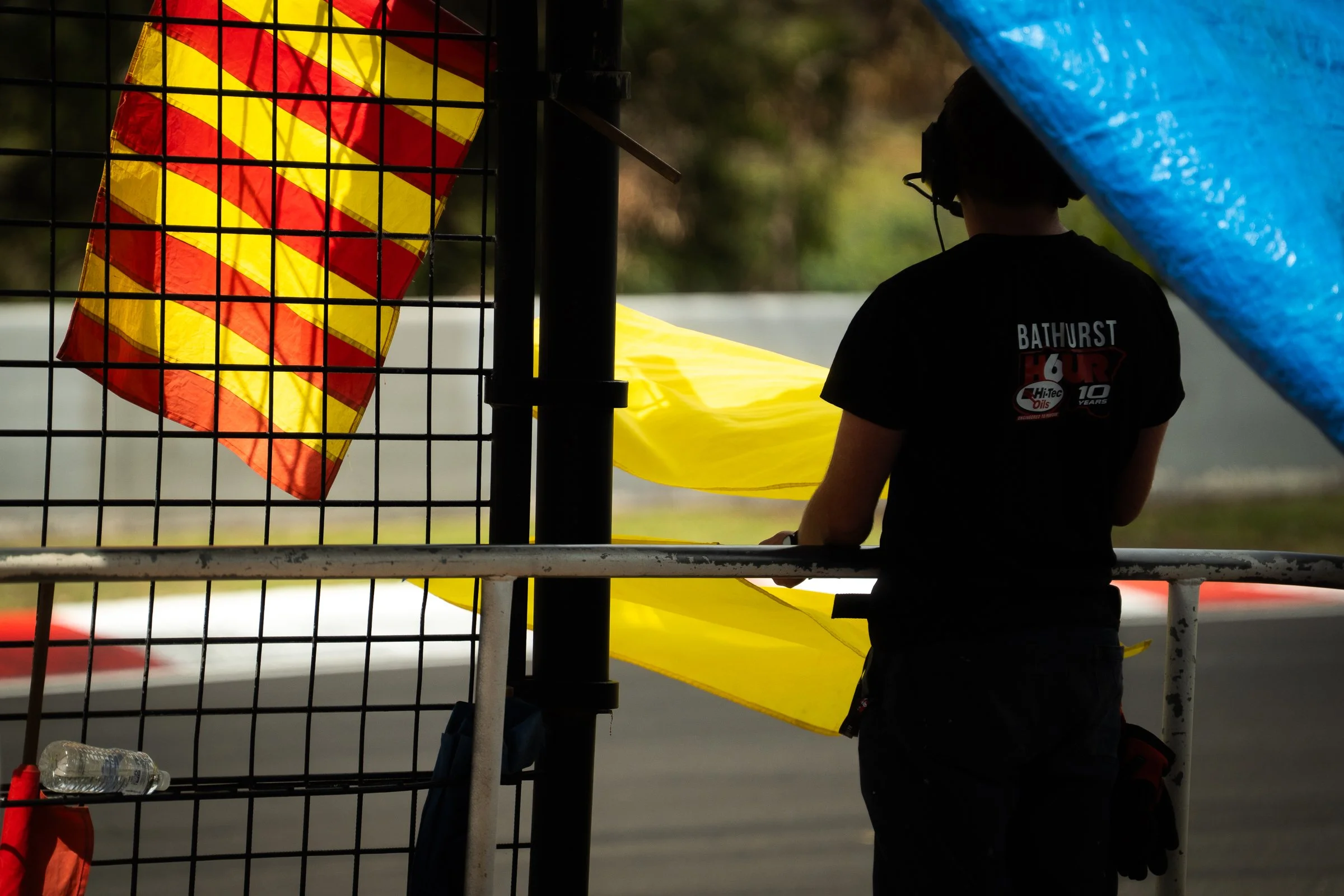 Person standing beside a racetrack barrier holding a yellow and red striped flag and a yellow flag under a blue tarpaulin