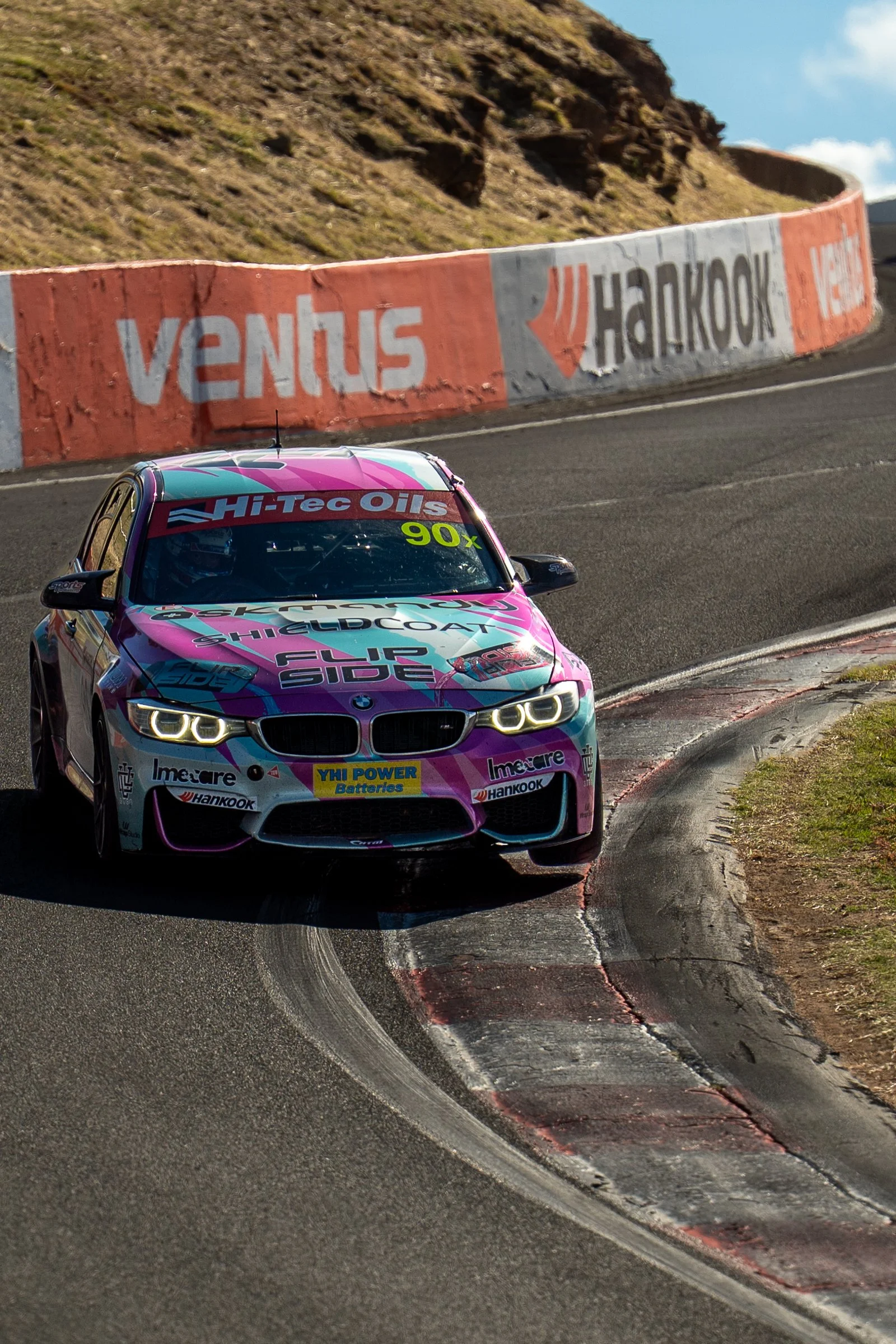 Front view of the Flip Side Racing BMW on track, turning into a corner over a red and white kerb, with its pink, blue and white livery and sponsor logos visible