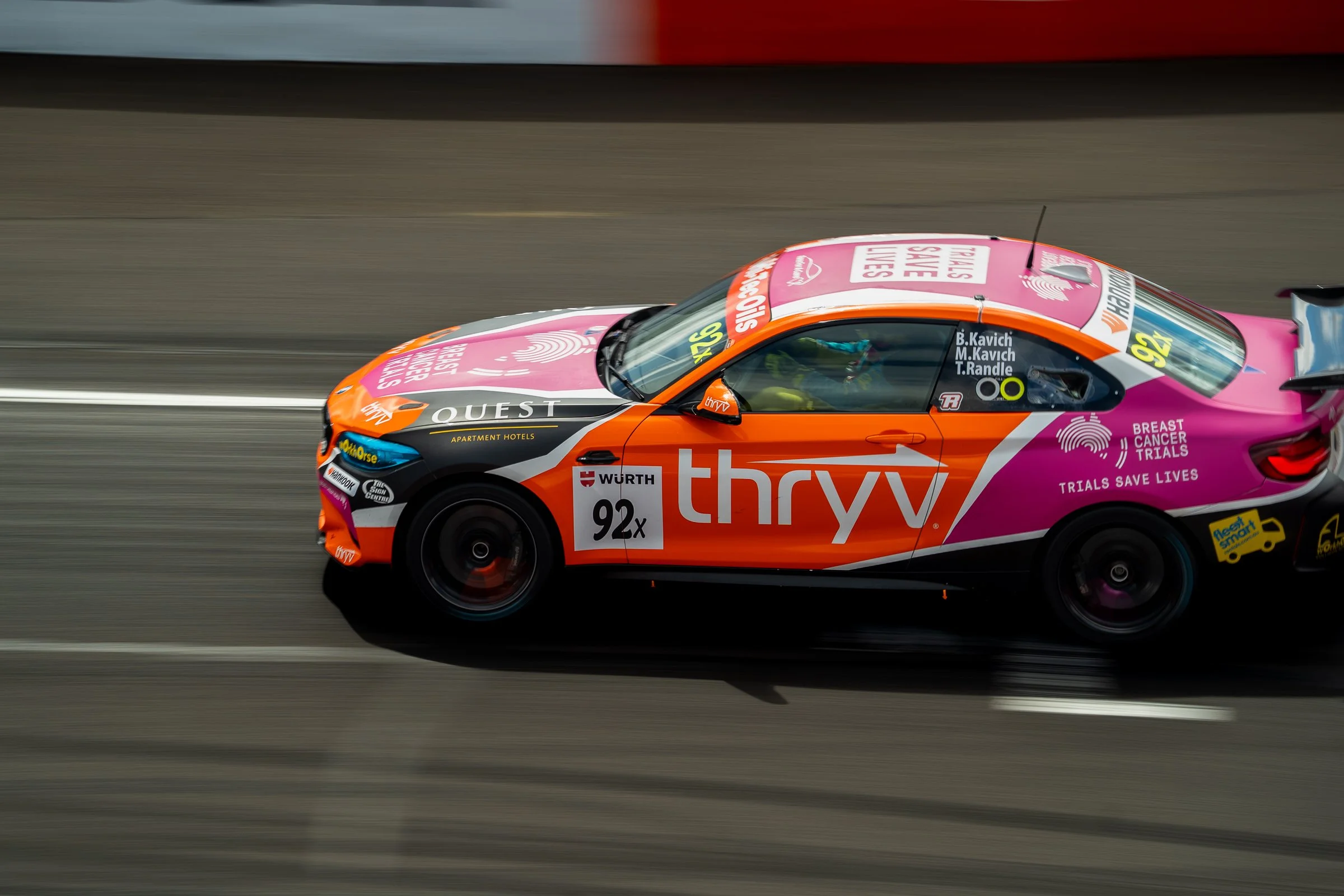 BMW race car with pink, orange and black livery racing on track, showing Thryv and Breast Cancer Trials branding with driver names visible on the side window