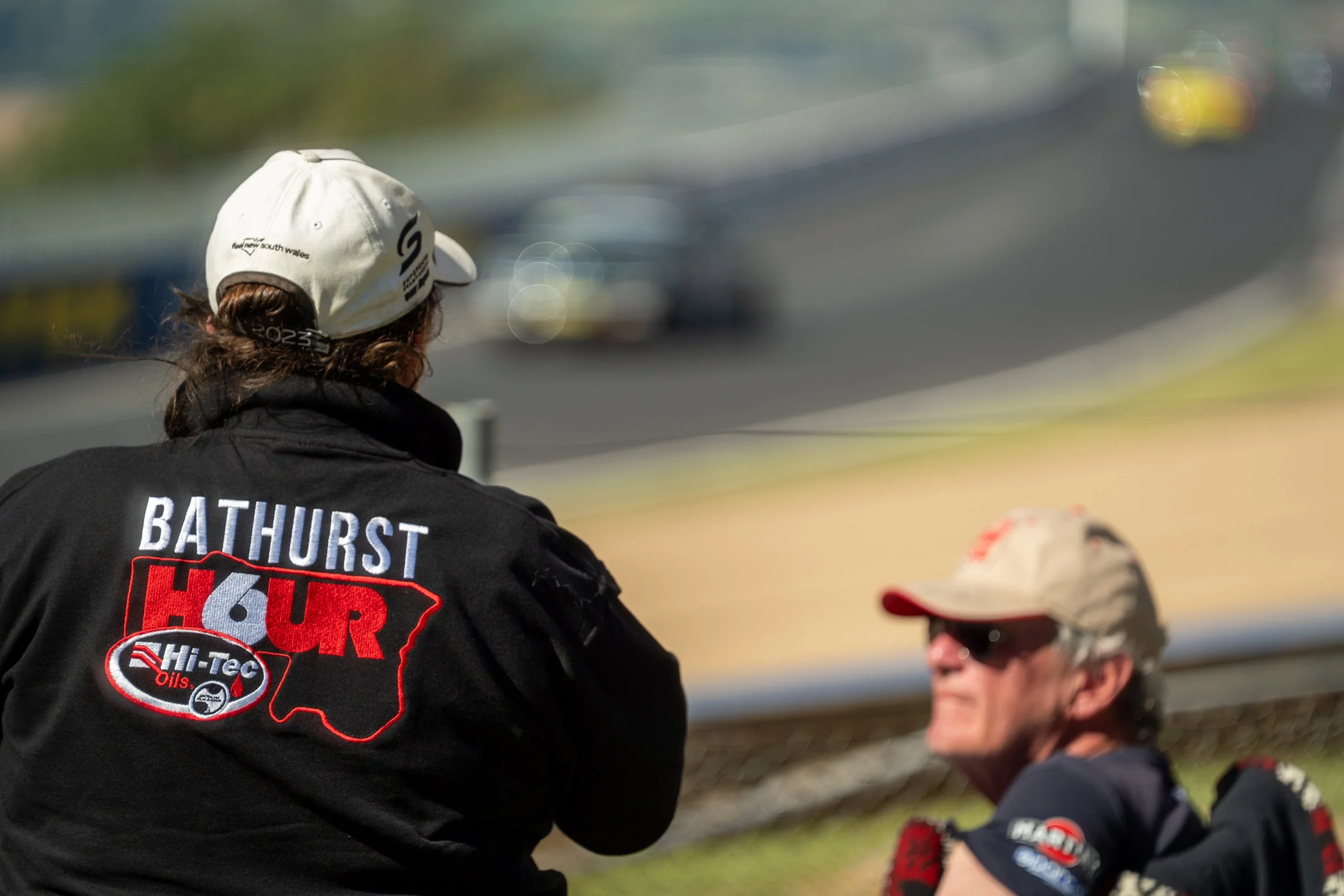 Two spectators sitting on the grass at a racing event, one wearing a Bathurst 6 Hour jacket, with race cars blurred in the background
