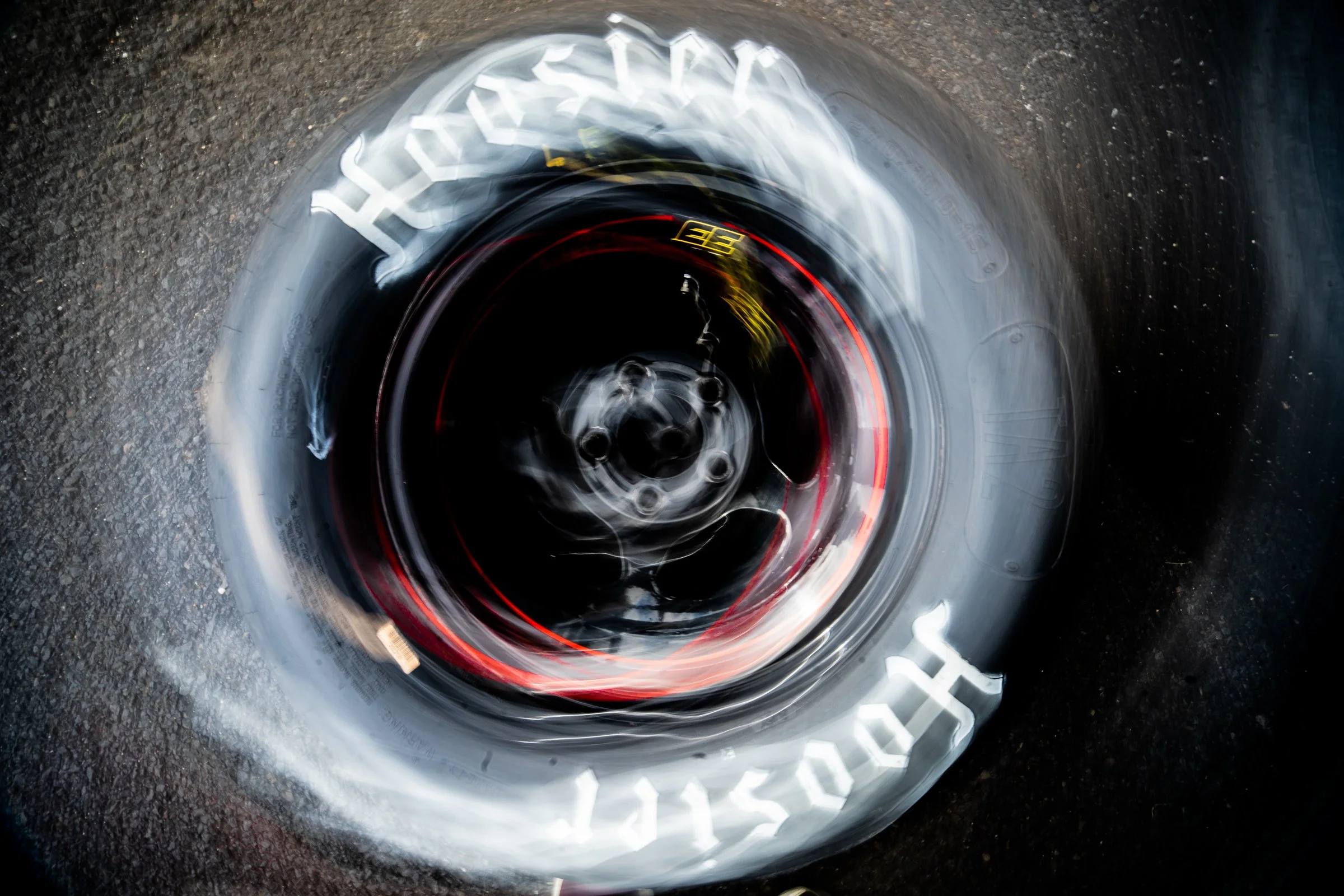 Close‑up of a Hoosier racing tyre with radial motion blur creating a spinning circular effect against the asphalt surface
