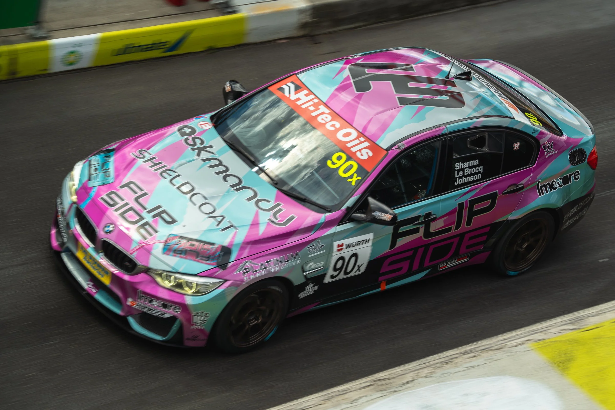 Overhead view of a race car with pink, blue and black livery speeding on track, with Flip Side branding and driver names visible on the window