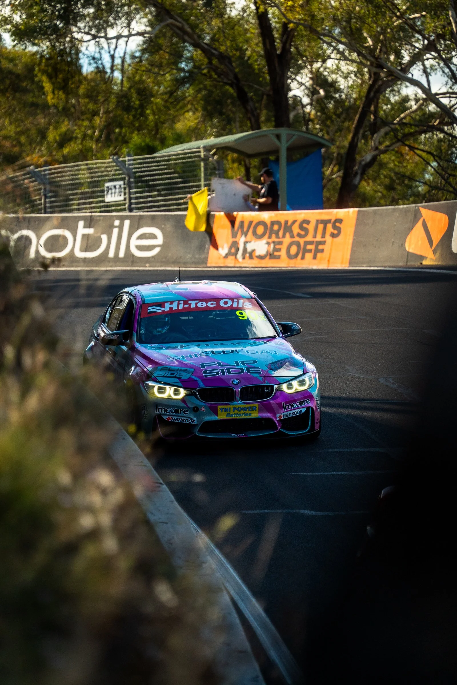Side Flip BMW race car driving downhill on track with headlights illuminated, framed by a tree and barrier in the foreground