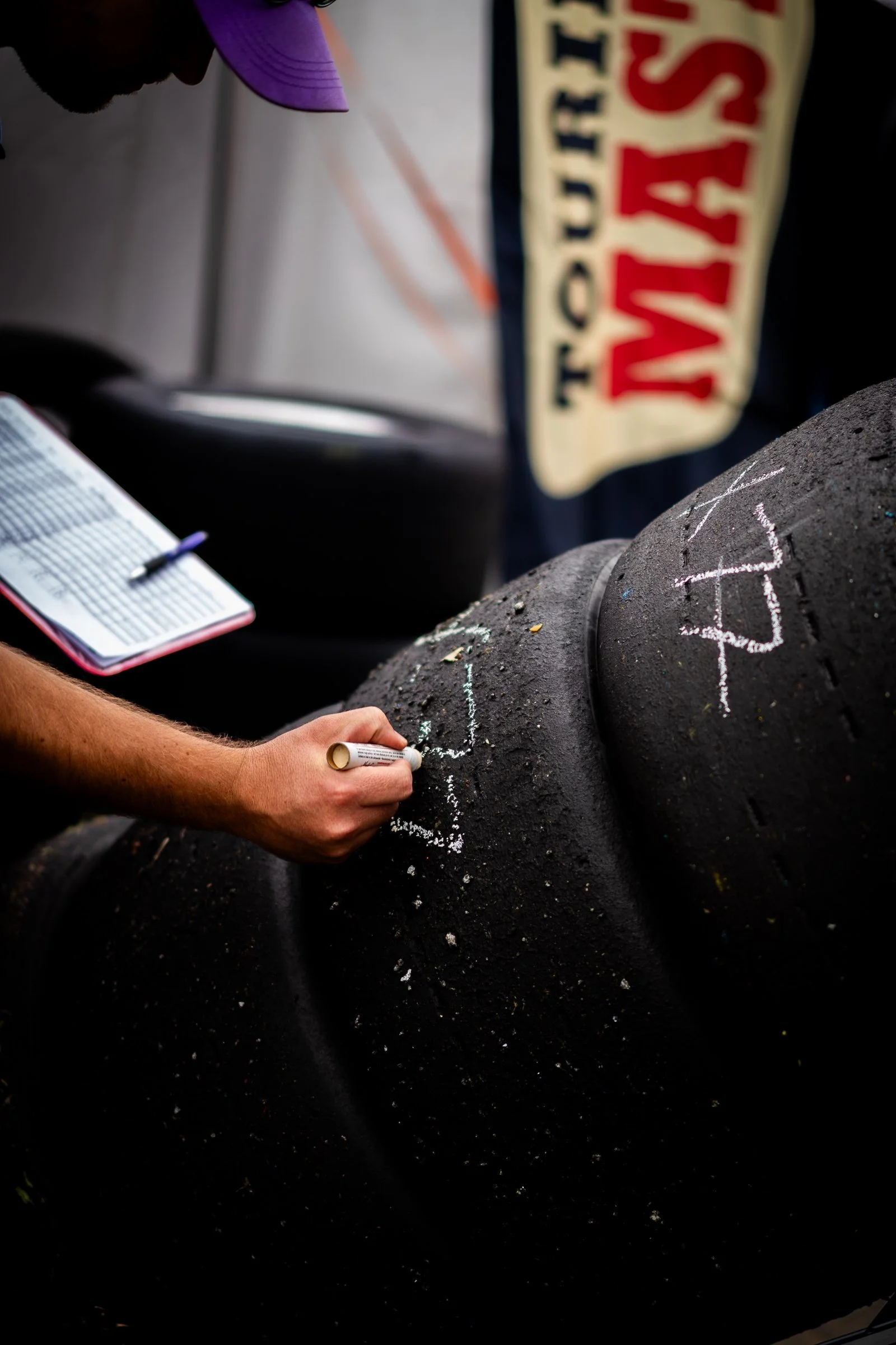 Person writing numbers in chalk on black racing tyres with a notebook and flag in the background
