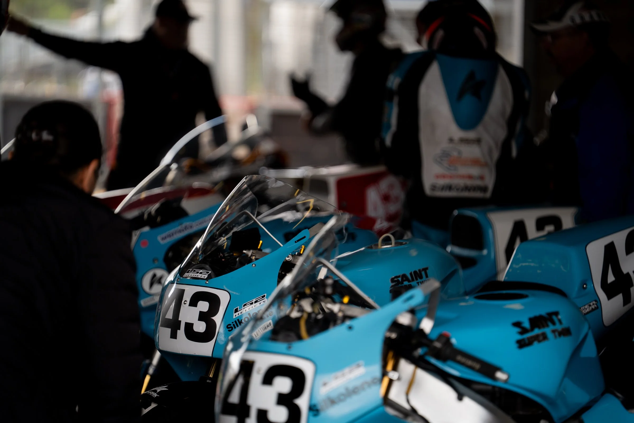 Group of blue racing motorcycles with number 43 being prepared by team members inside a garage