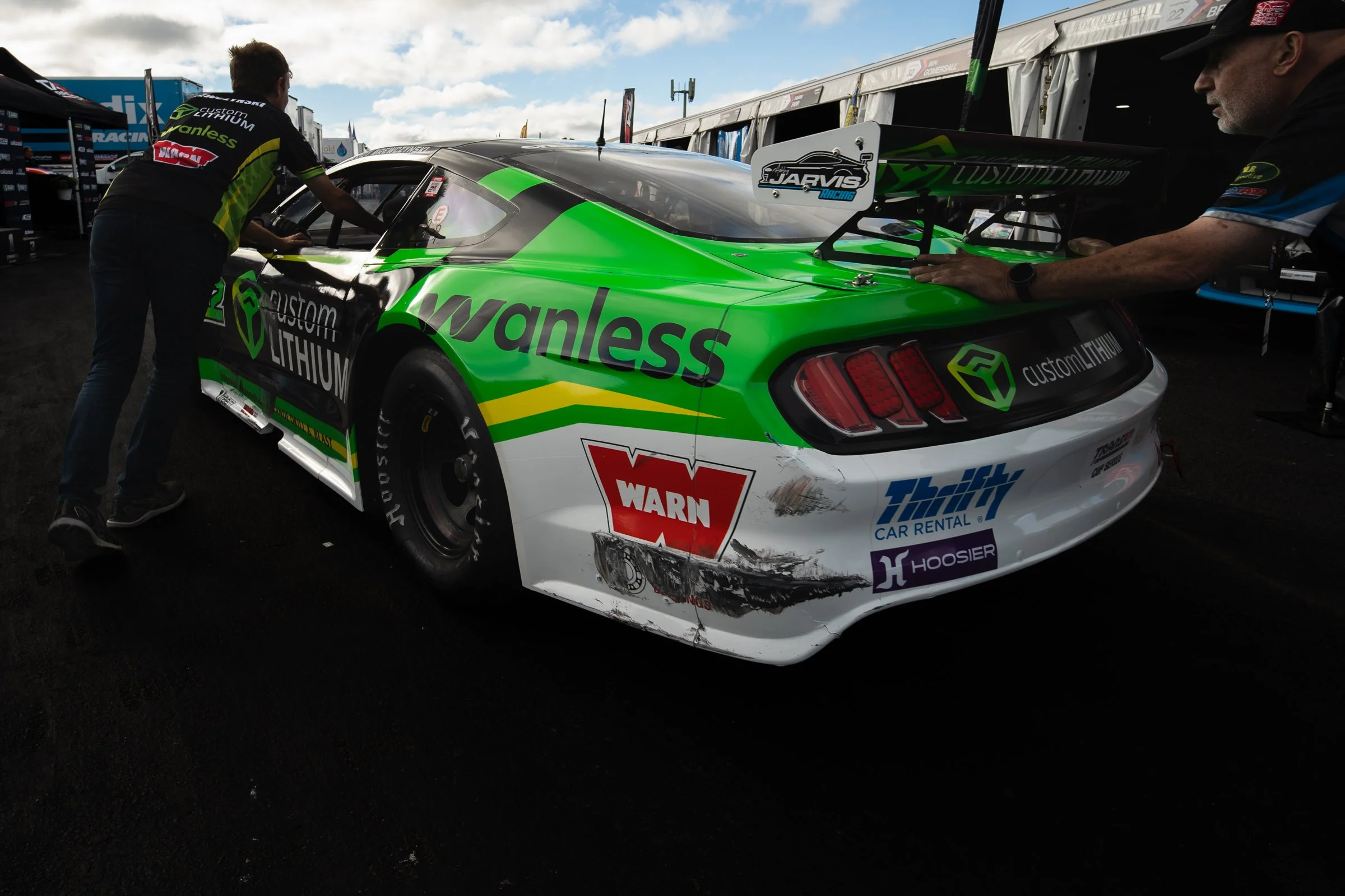 Green, black and white race car in a paddock area being pushed by two mechanics, with sponsor logos visible