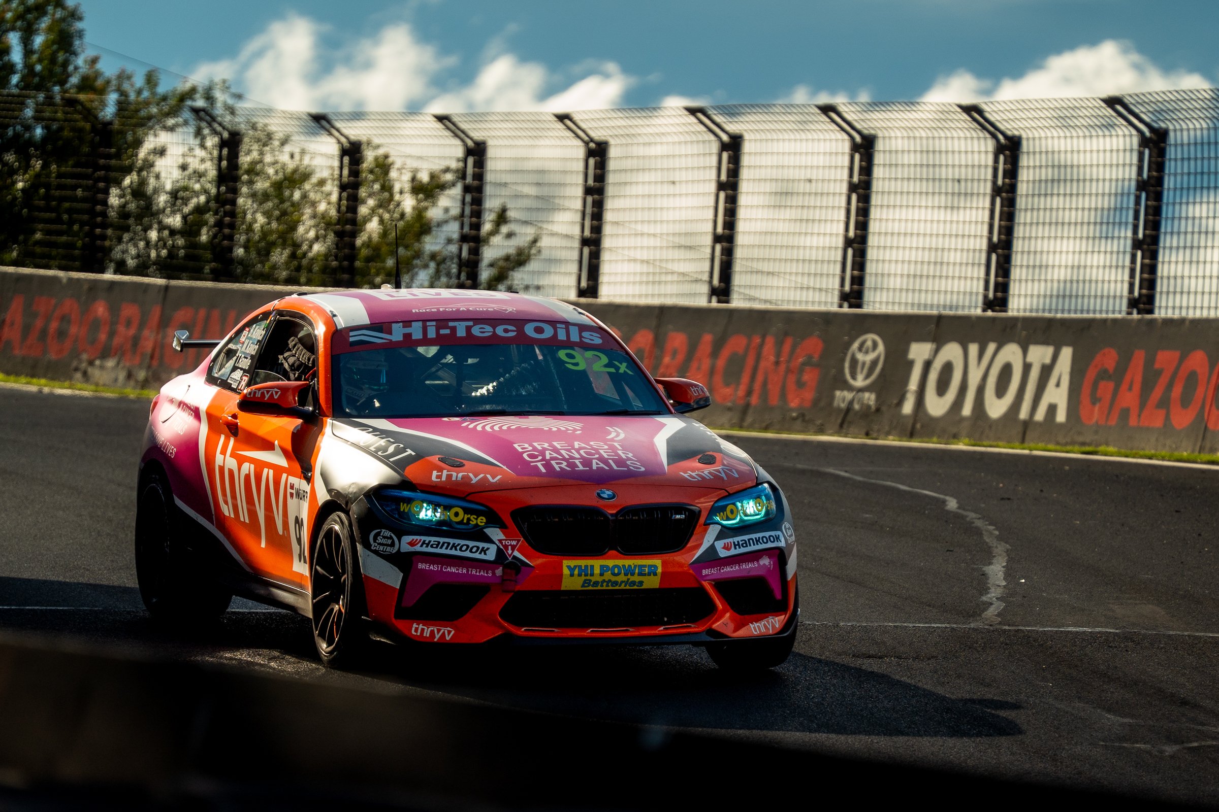 Pink and black BMW race car with sponsor logos navigating a turn on the racetrack with barriers and advertisements in the background