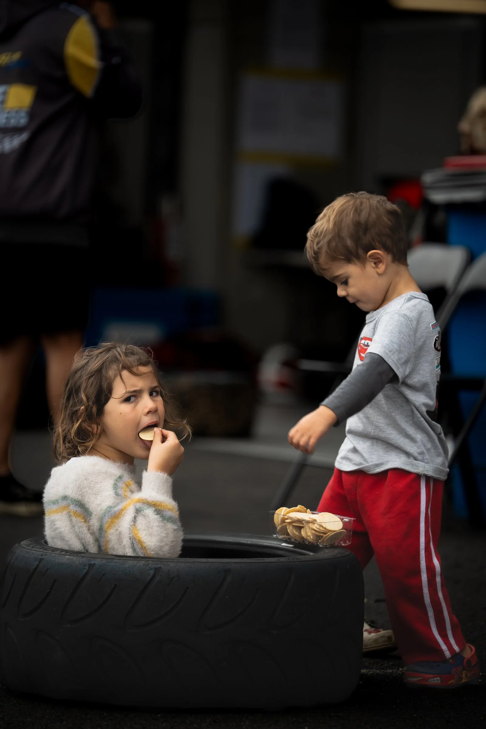 Young girl sitting inside a race car tyre eating a biscuit from a tray resting on the tyre, with a young boy standing nearby