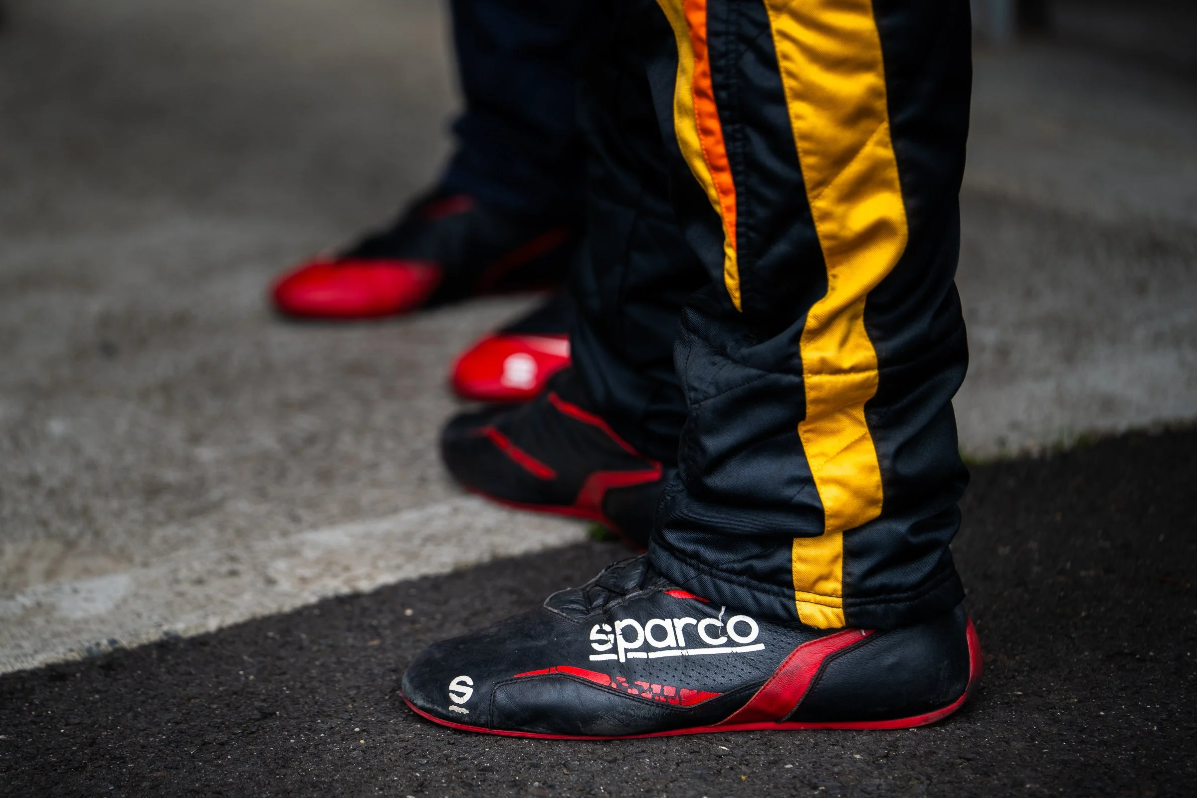 Close‑up of a race driver’s feet in racing shoes on the track surface, with another driver’s feet visible in the background