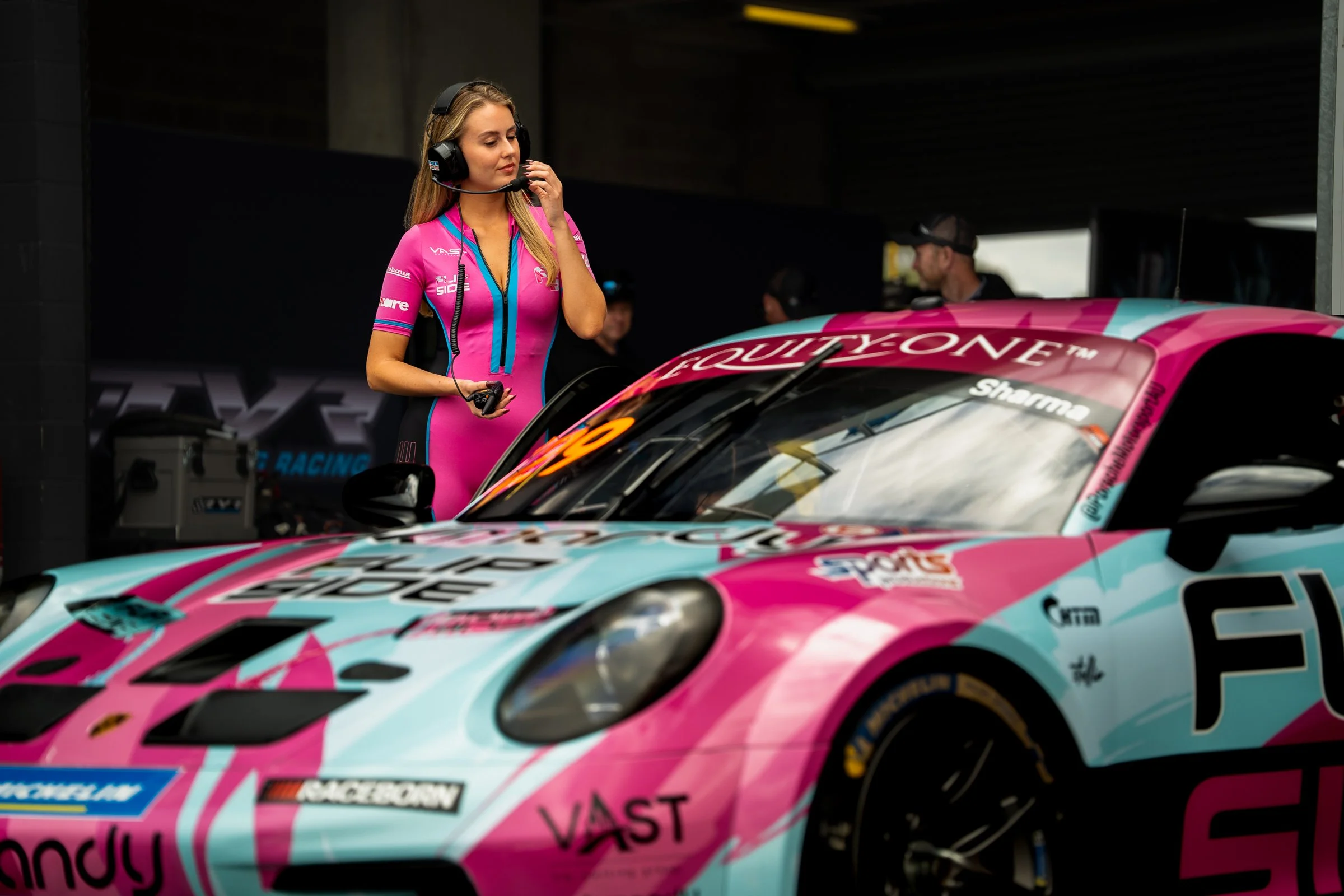 Promotional model in a pink and blue bodysuit wearing a headset standing near a pink and blue Porsche race car in front of a garage