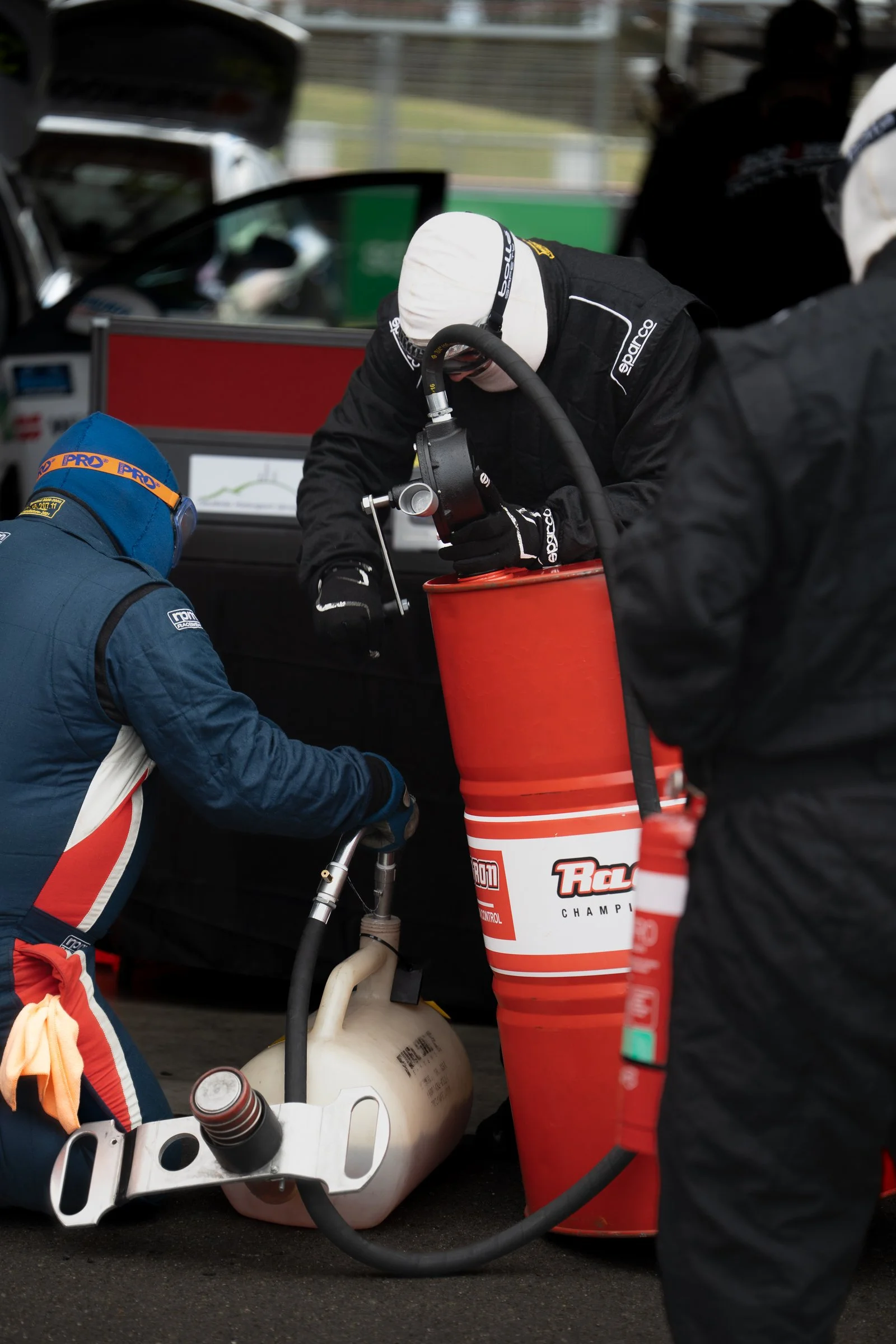 Pit crew members filling their refuelling rig from a large fuel drum using a hand‑crank transfer pump and black hose