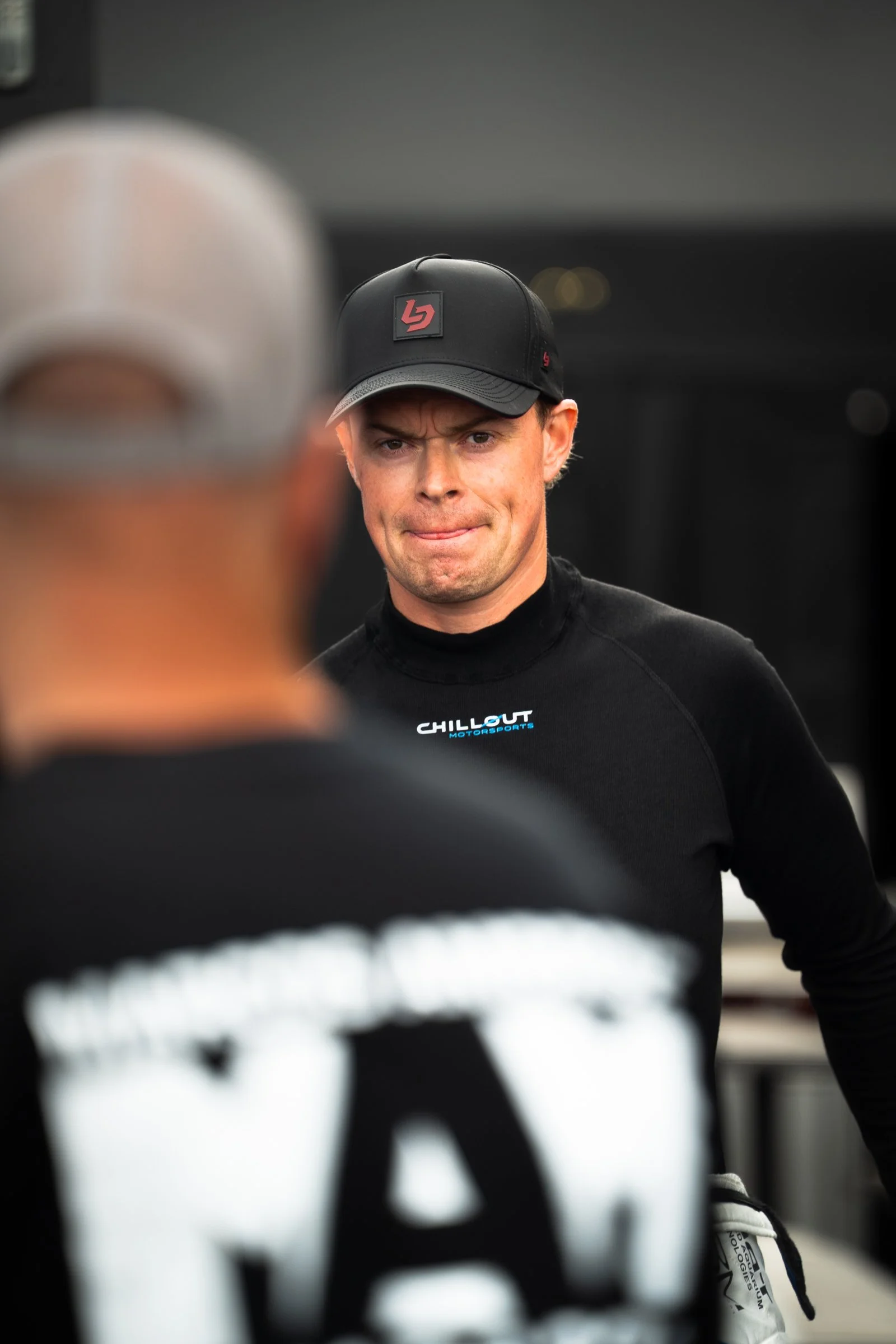 Person wearing a black cap and a Chillout Motorsports shirt standing in a garage‑area setting, looking toward another person in the foreground