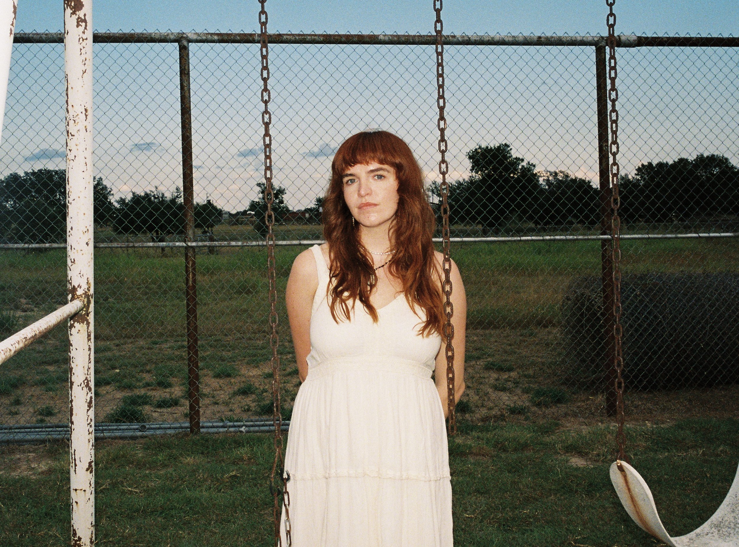 girl with bangs and red hair in a white vintage dress on an old swingset in front of a chain link fence that's rusty