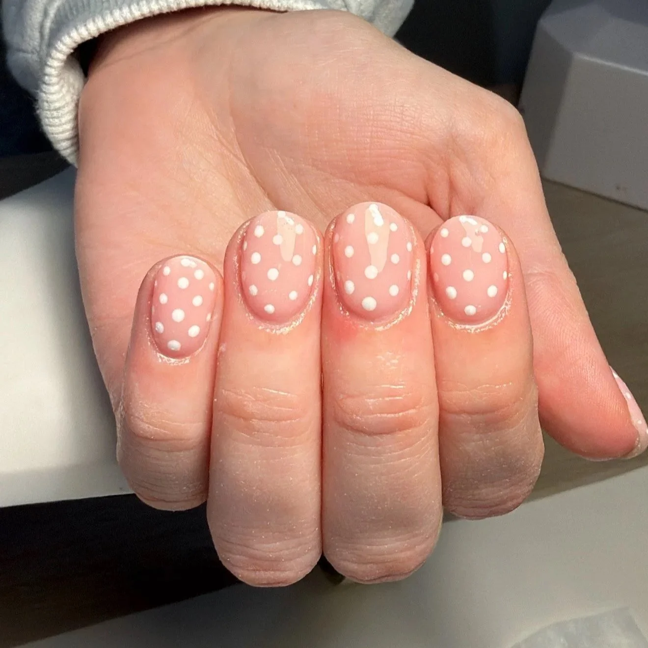 Close-up of a hand with oval-shaped nails painted with pink nail polish and white polka dots.