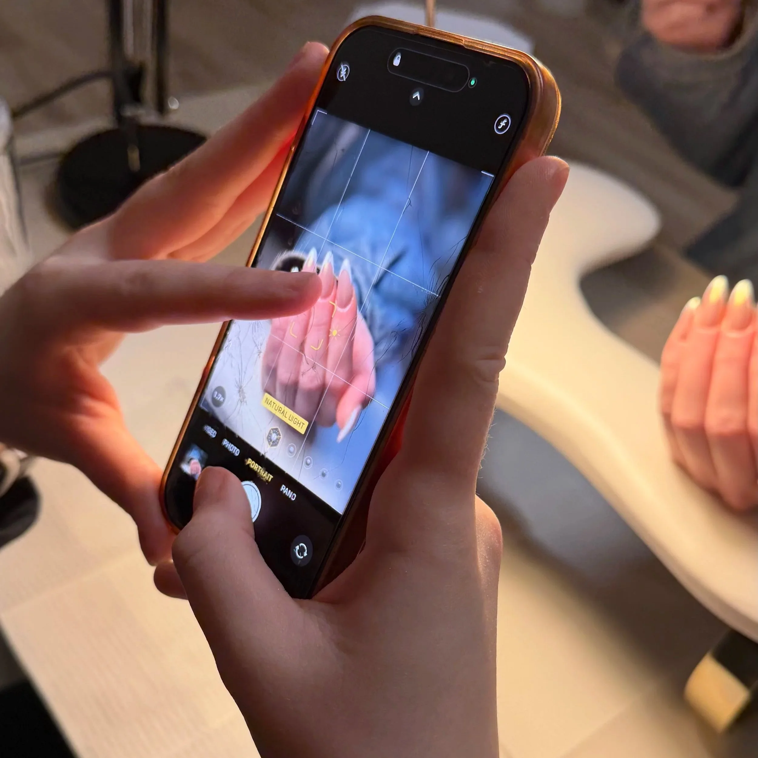 Person taking a photo of another person's hand with their smartphone, focusing on the hand with long manicured nails, in a setting with soft lighting.