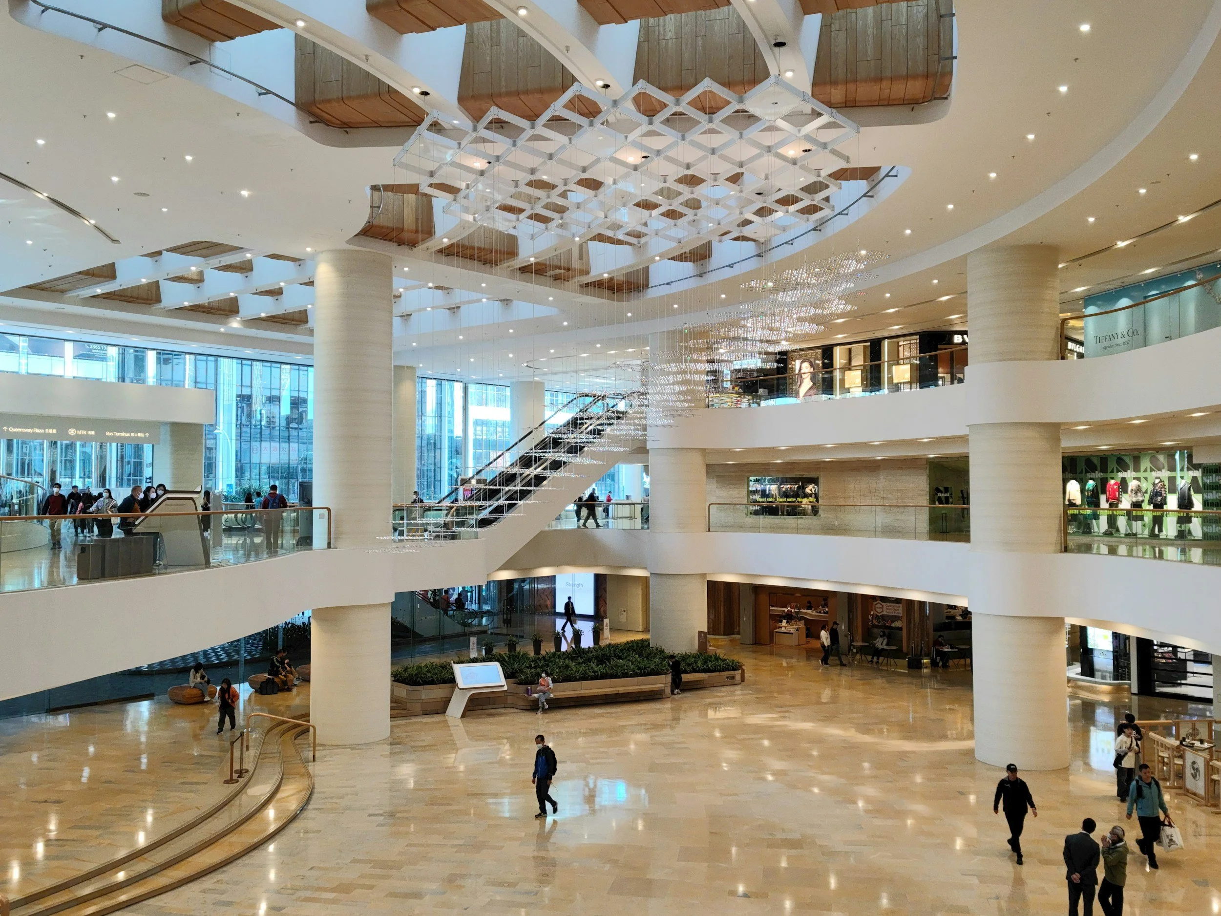 Interior view of a multi-level shopping mall with escalators, glass storefronts, and shoppers walking on the polished marble floors.