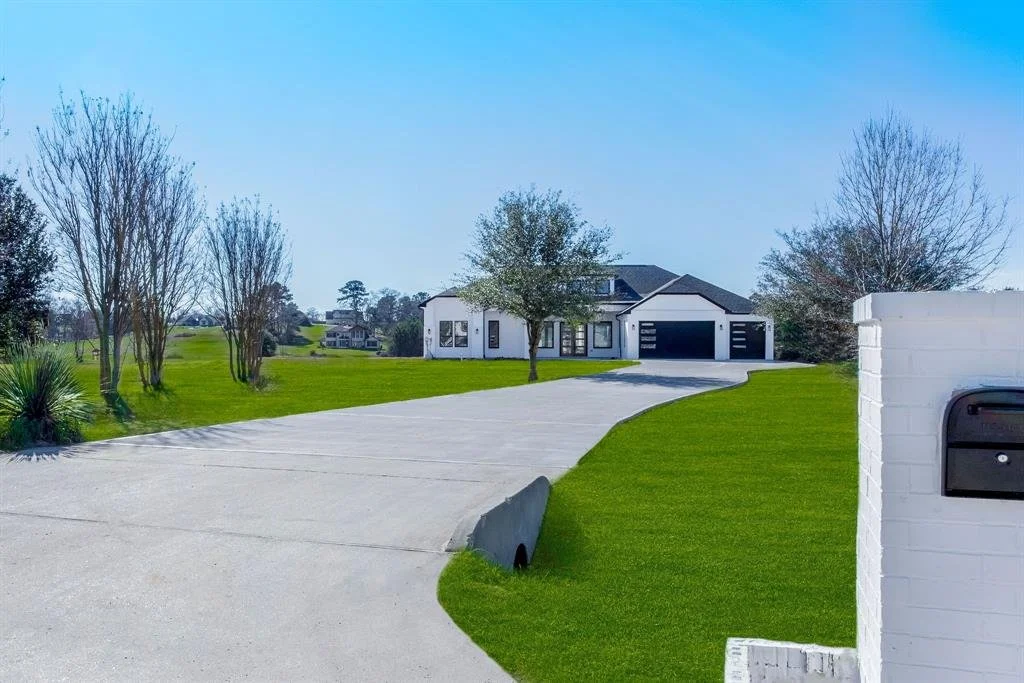 A modern house with a driveway, surrounded by green grass and trees, with a white brick wall and mailbox in the foreground.