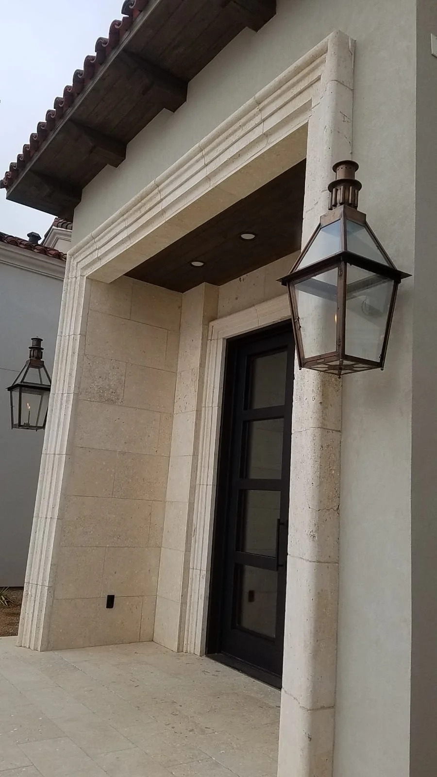Exterior view of a building entrance with black door, stone frame, and two metal lantern-style light fixtures.