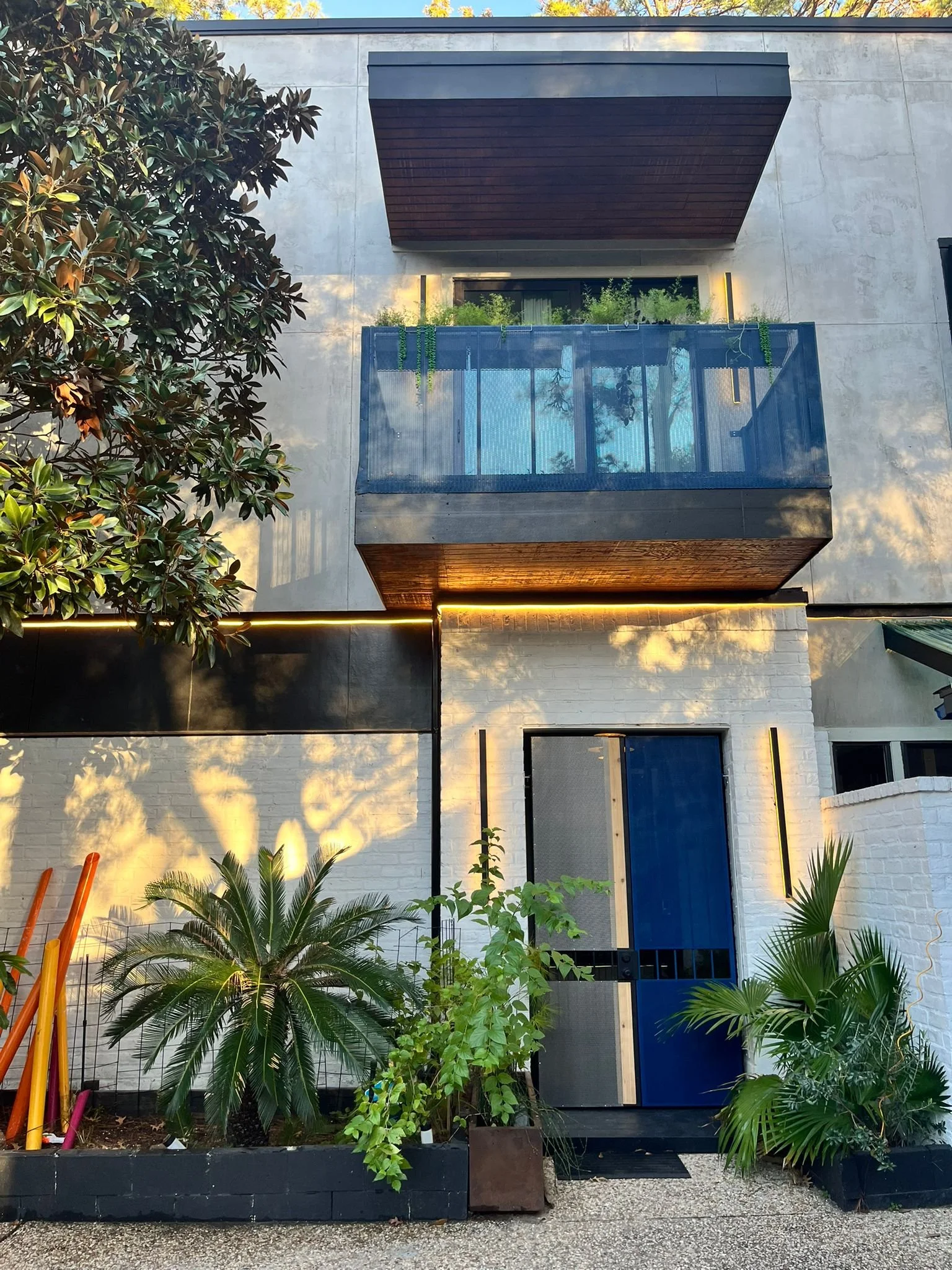 Exterior of a modern multi-story building with a small garden in front, featuring palm plants, a blue door, and a balcony with greenery. Modern wall lights and a clean, minimalist design.
