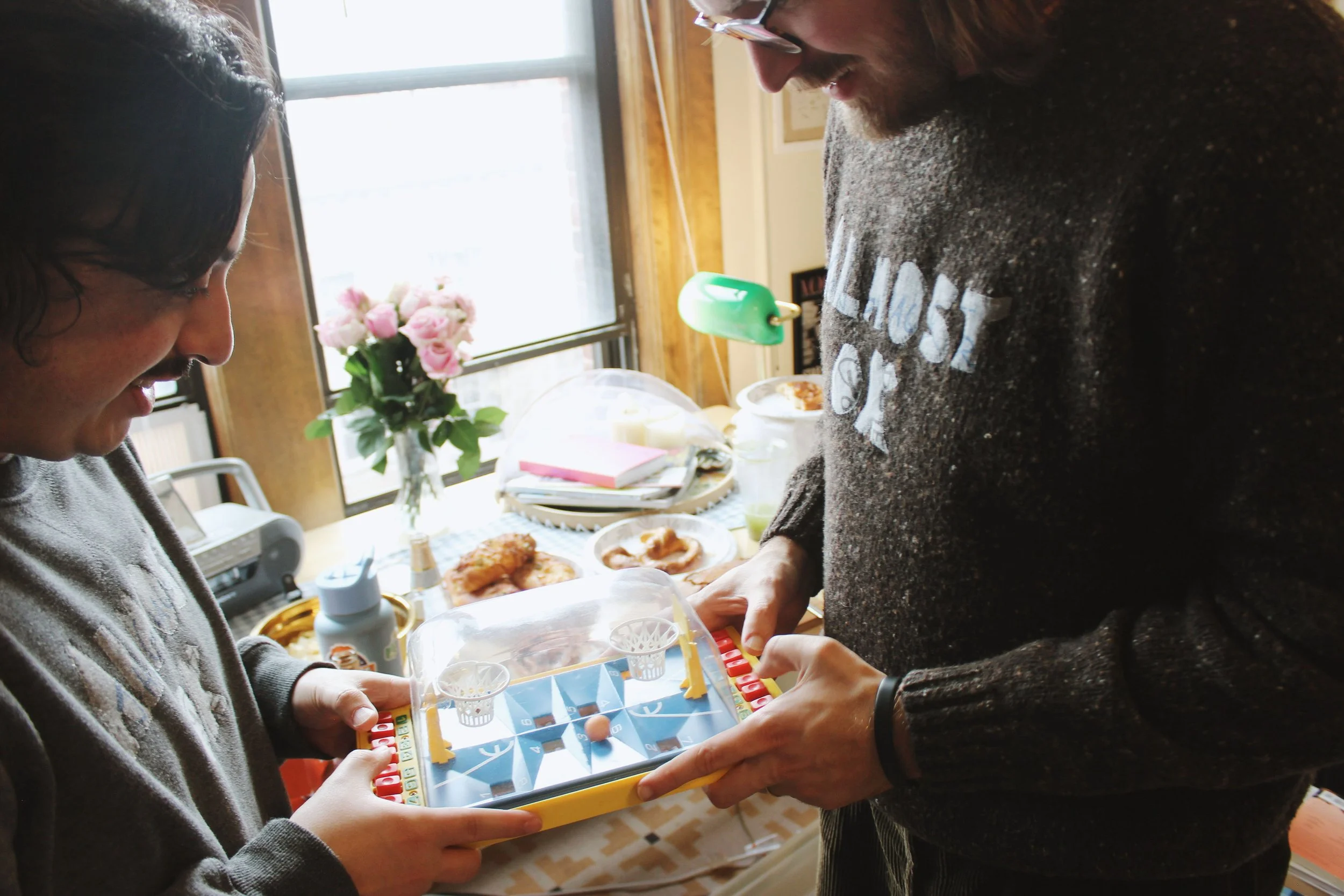Two people playing a tabletop hockey game in a cozy, well-lit room with a window, flowers, and snacks in the background. They are wearing sweaters with patches.