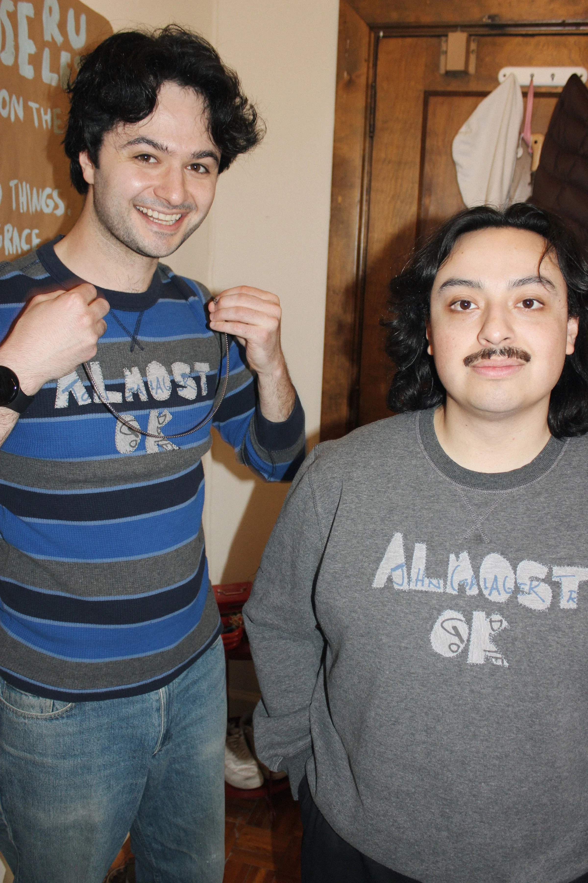 Two men standing indoors, one smiling and holding a necklace, the other with a neutral expression. Both are wearing casual clothing, with one in a striped shirt and the other in a gray sweatshirt. Background includes a wooden door, hooks with hanging