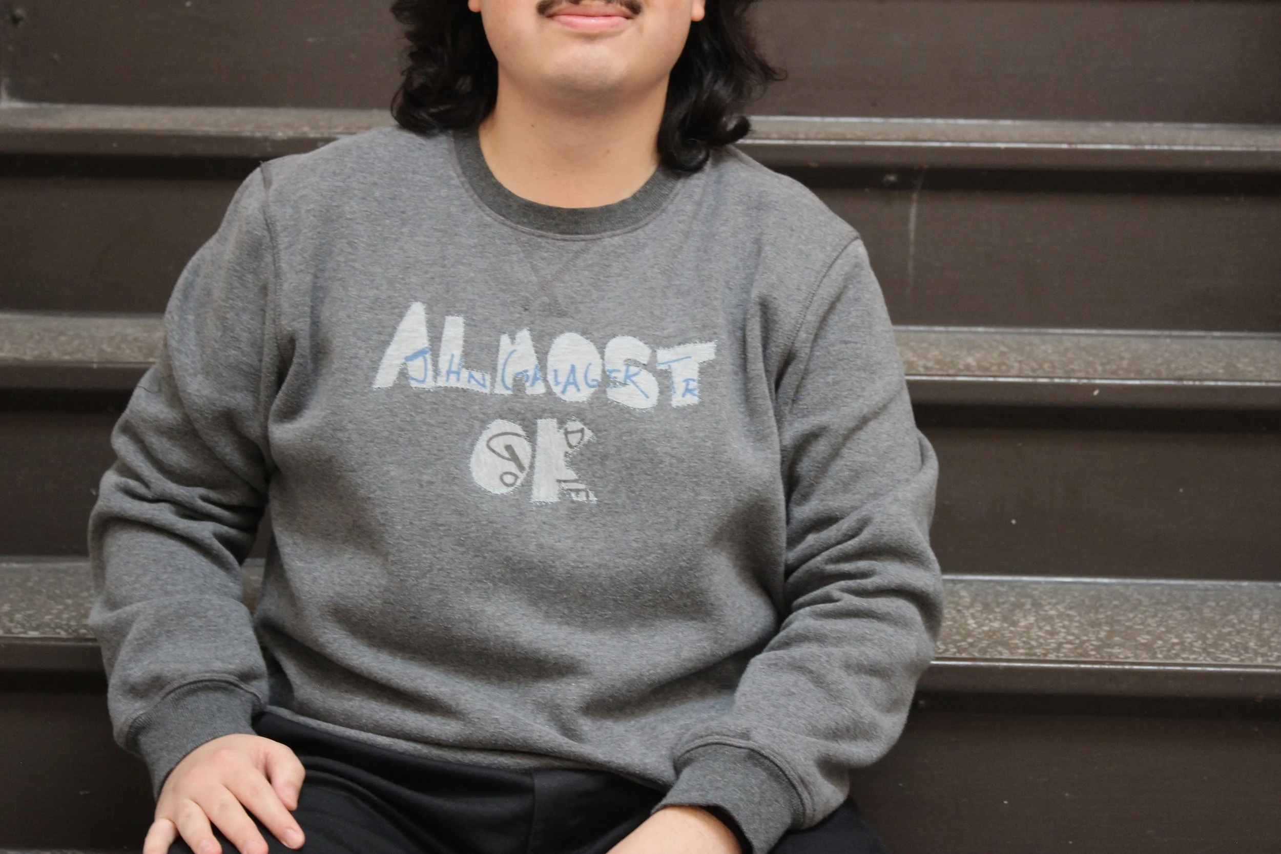 Person sitting on stairs wearing a gray sweatshirt with text patches.