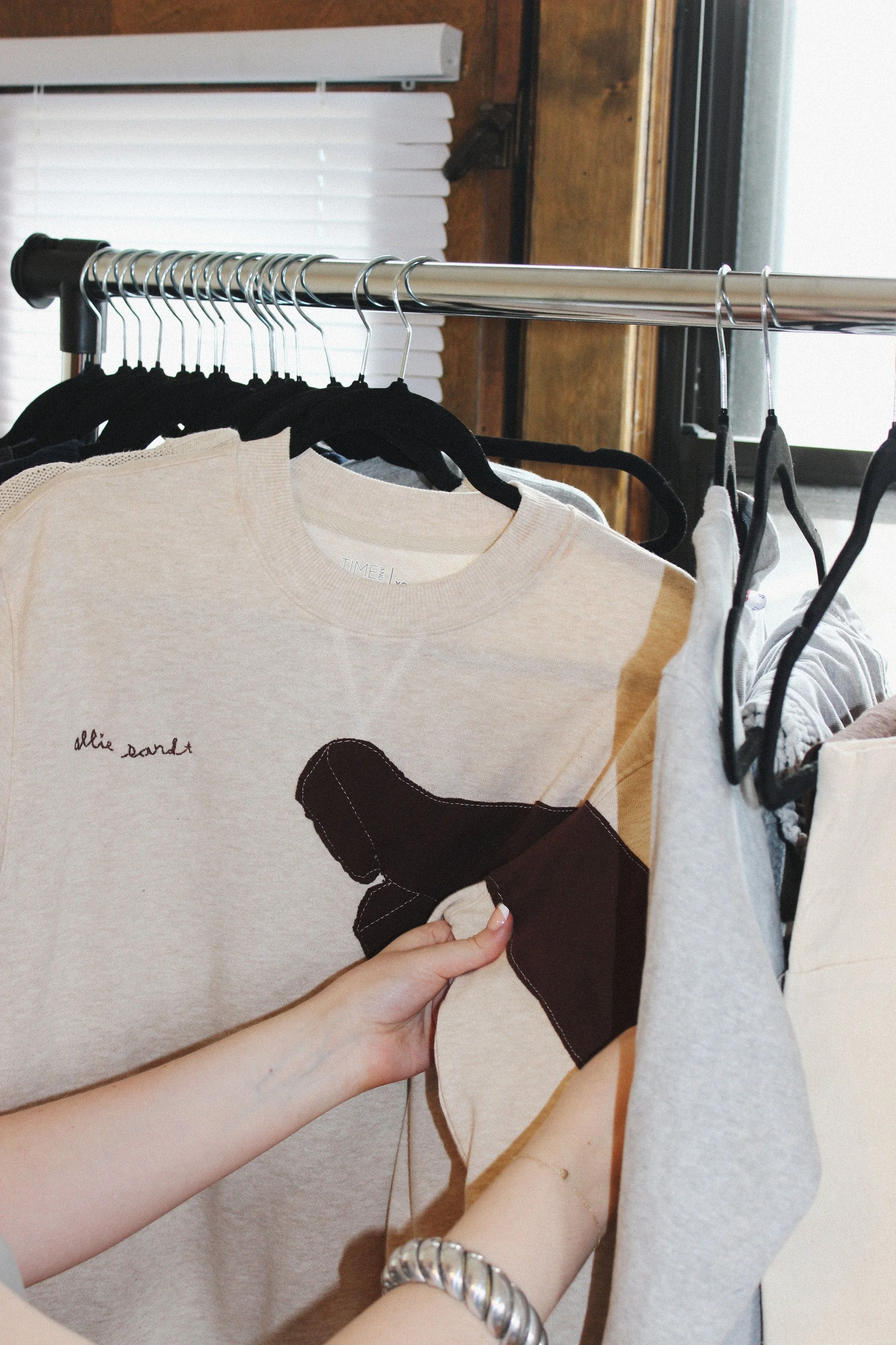 A person holding a beige patchwork T-shirt with a silhouette of a boot on it, hanging on a clothing rack among black and gray shirts.
