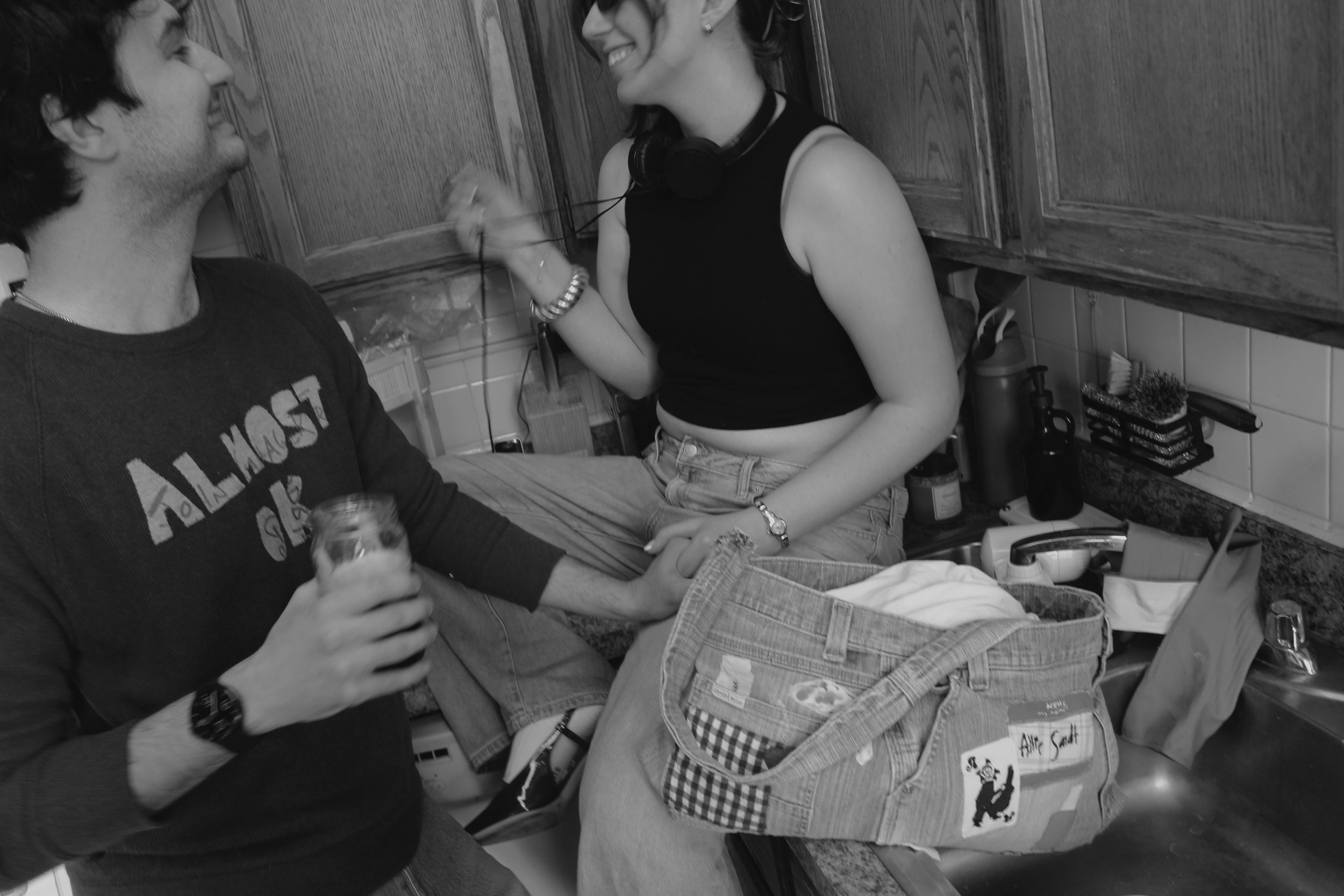 A man and woman in a kitchen, smiling and holding hands. The woman is sitting on the counter with a pair of headphones around her neck.