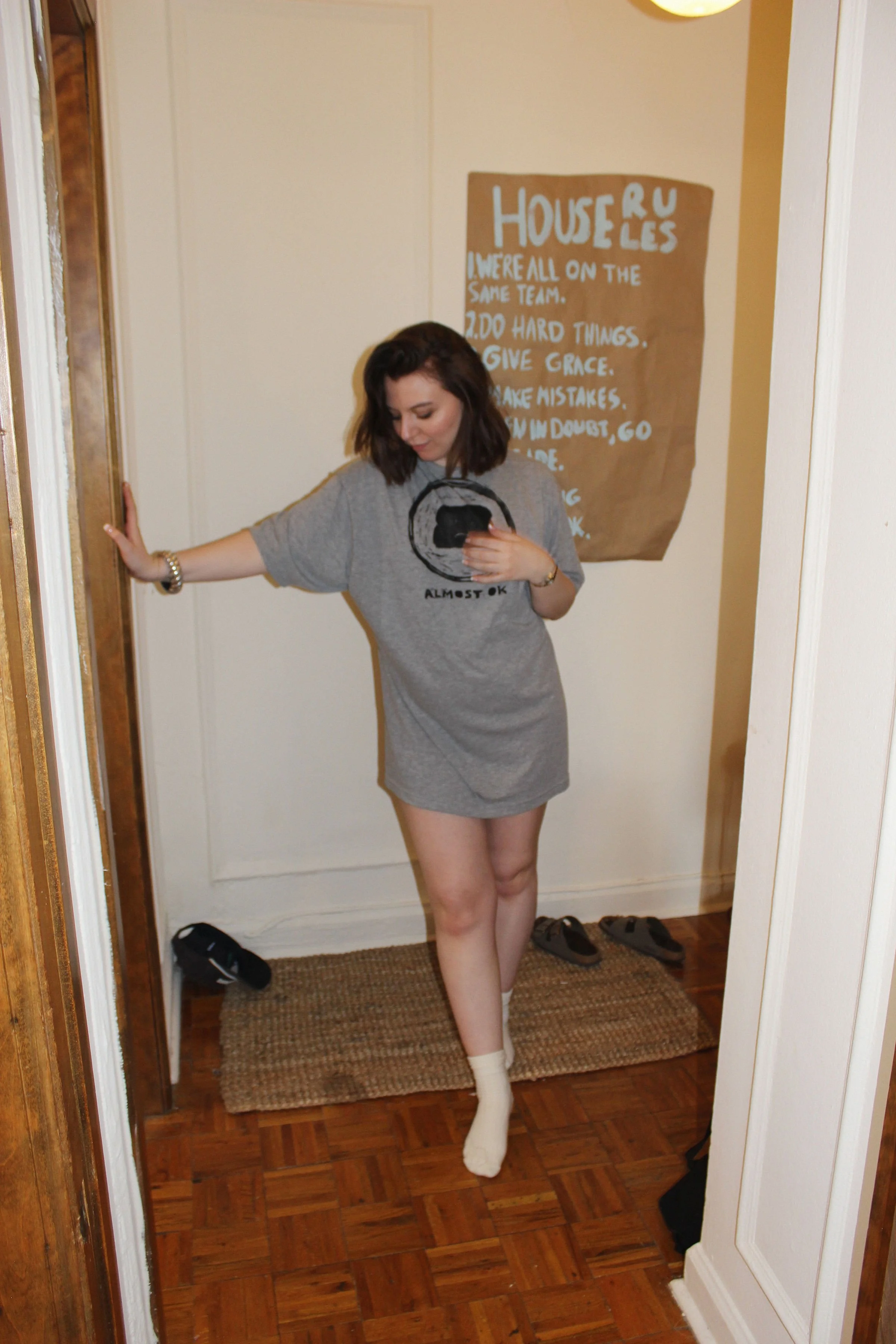 A woman in a gray oversized block printed T-shirt and socks, taking a selfie in a mirror in a hallway. Behind her is a brown paper with handwritten notes on the wall.