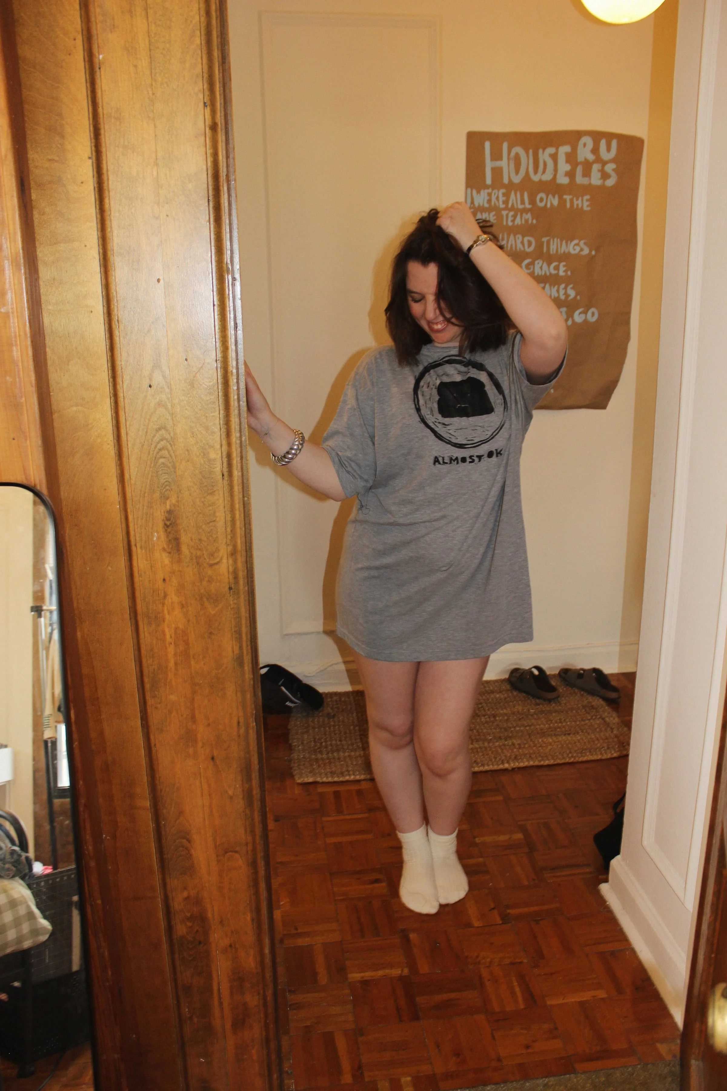 A woman with short dark hair is standing in a doorway, wearing an oversized gray T-shirt with a hand printed graphic, white socks, and smiling while touching her hair.