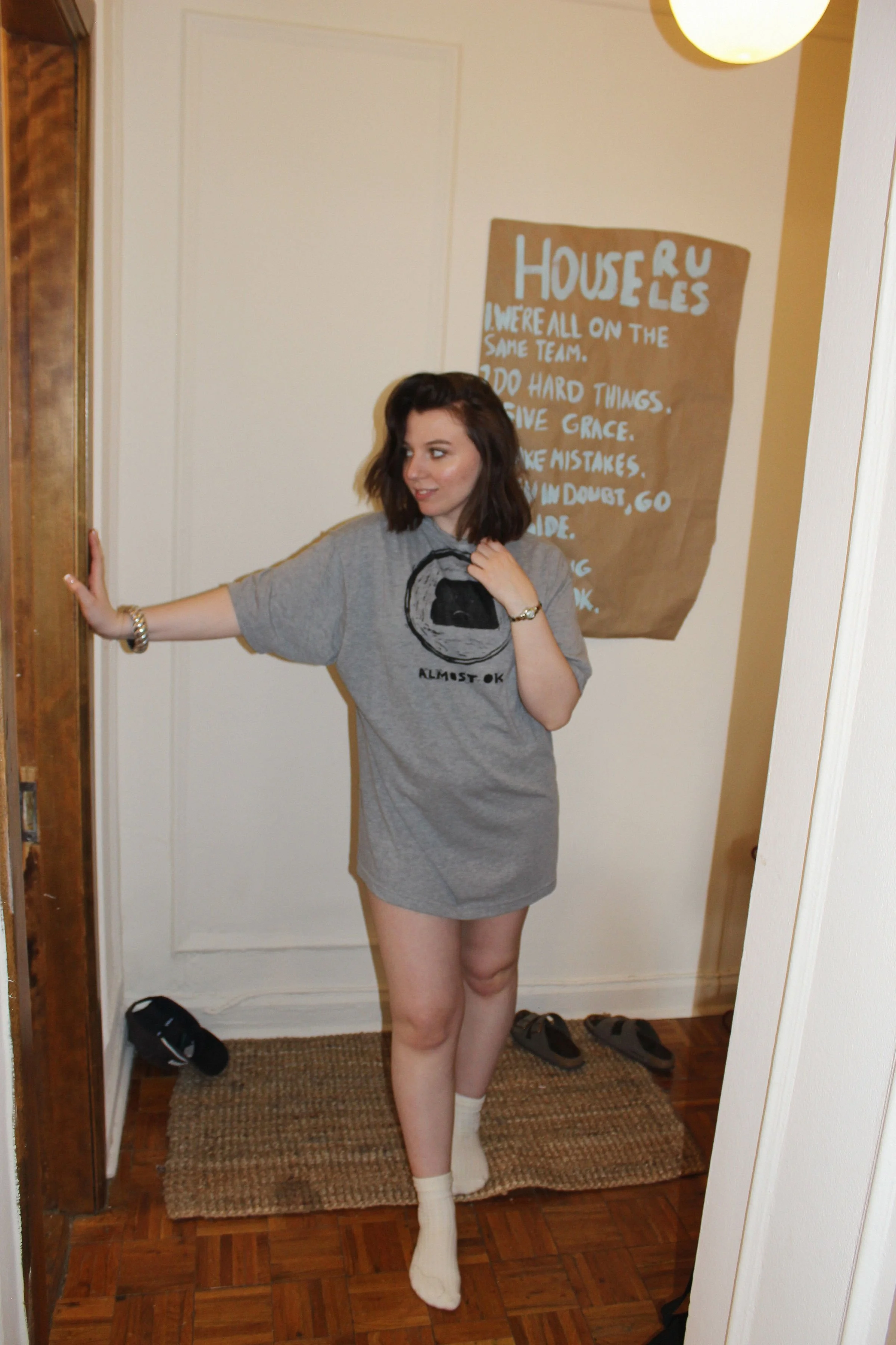 A young woman with dark, short hair wearing an oversized gray T-shirt with a hand printed graphic, standing on a woven rug near a doorway, with a large brown poster with blue text behind her.