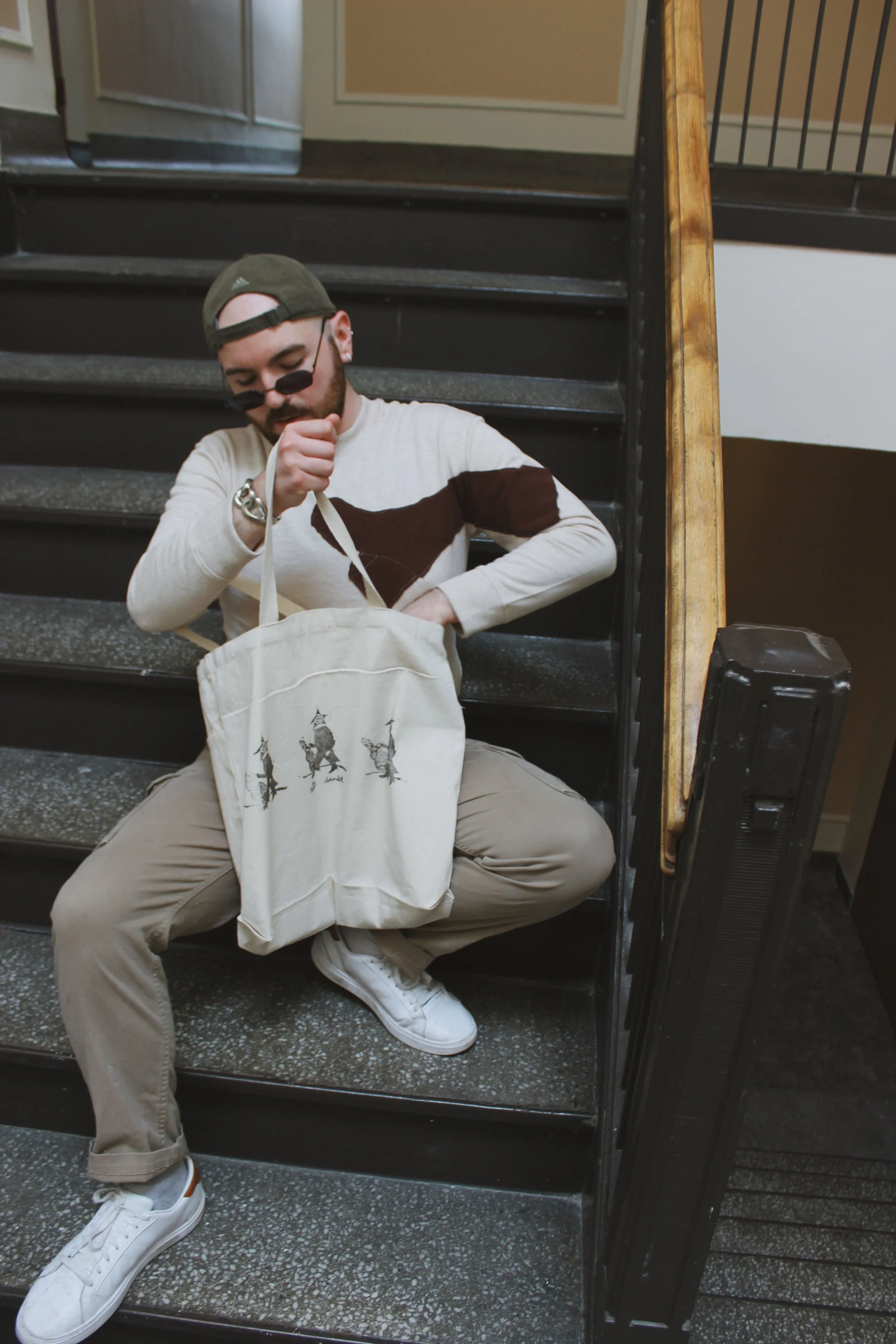 Man with glasses and cap sitting on stairs, looking inside a tote bag with horse illustrations, at indoor location.