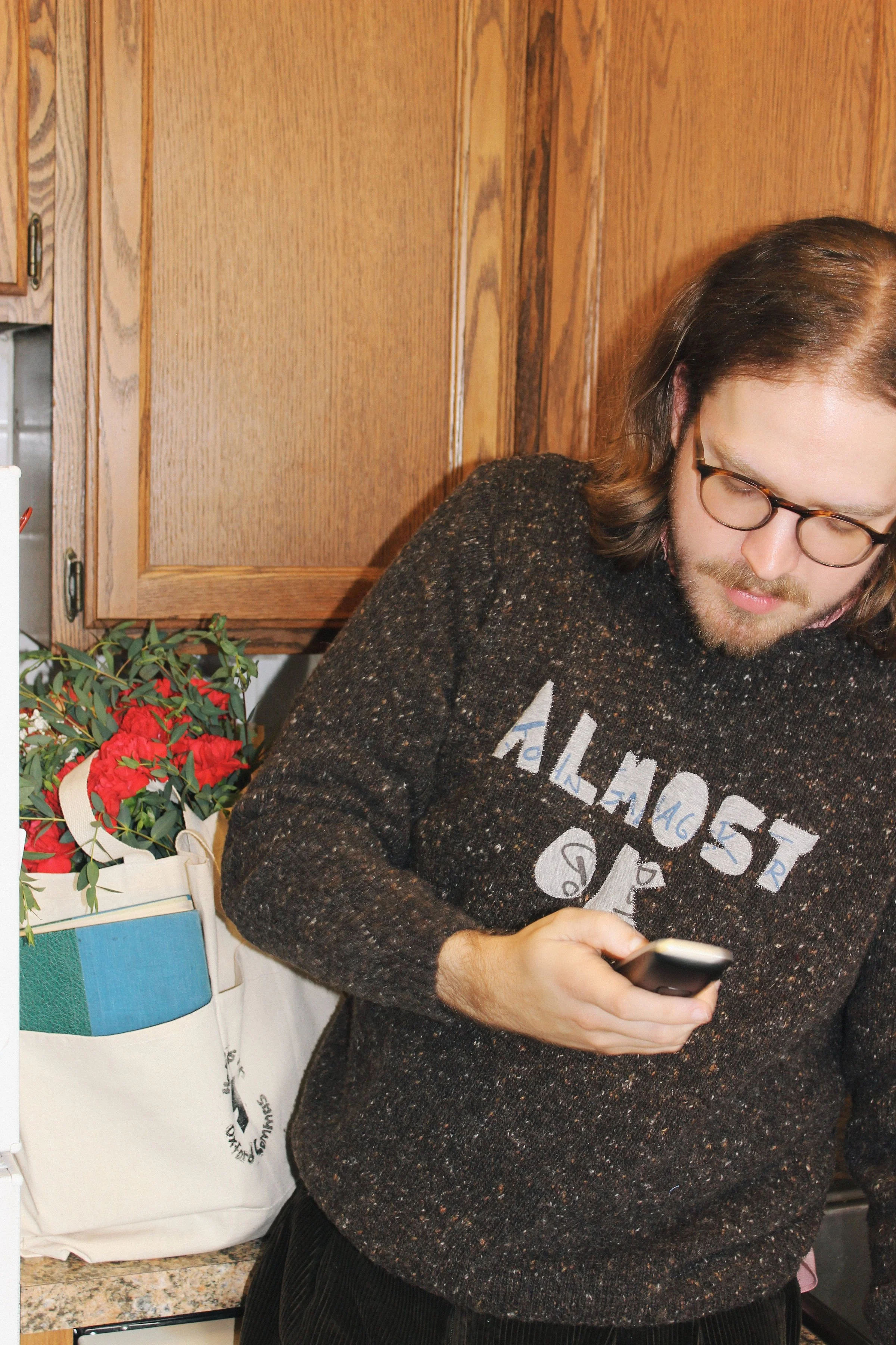 A man with glasses and shoulder-length hair looking down at his phone in a kitchen with wooden cabinets. There is an illustrated tote bag with flowers and books on the counter.