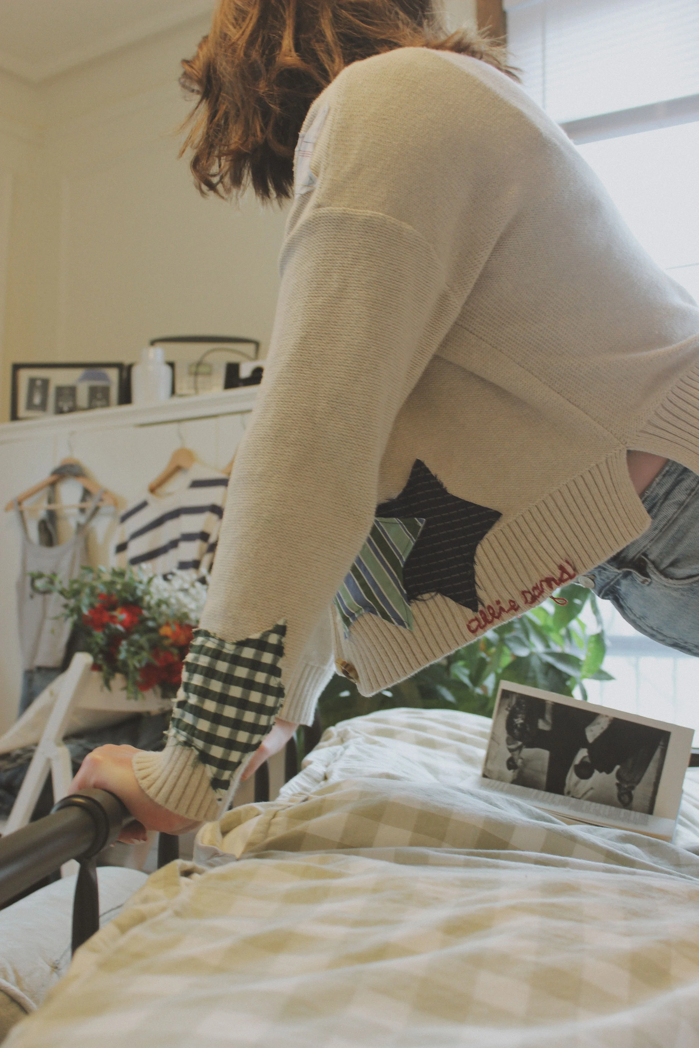 Person making the bed, seen from the side, with a focus on their hand and torso, wearing a beige sweater with patchwork details and the words 'allie sandt' embroidered, and jeans, in a cozy bedroom with and clothes hanging in the background.
