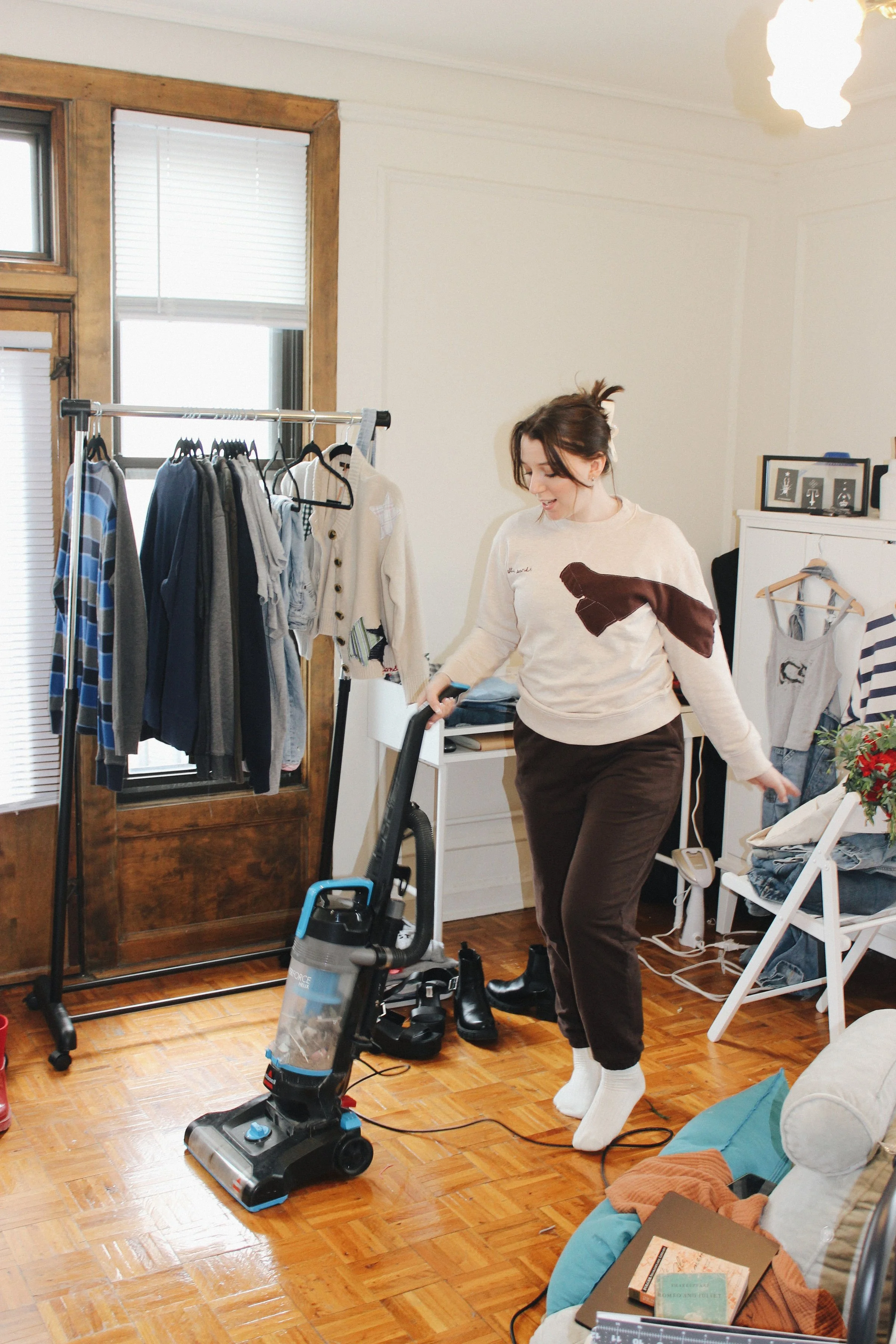A woman vacuuming a hardwood floor in a bedroom surrounded by clothing, shoes, and household items. She is wearing a music merch sweatshirt with patches and embroidery.
