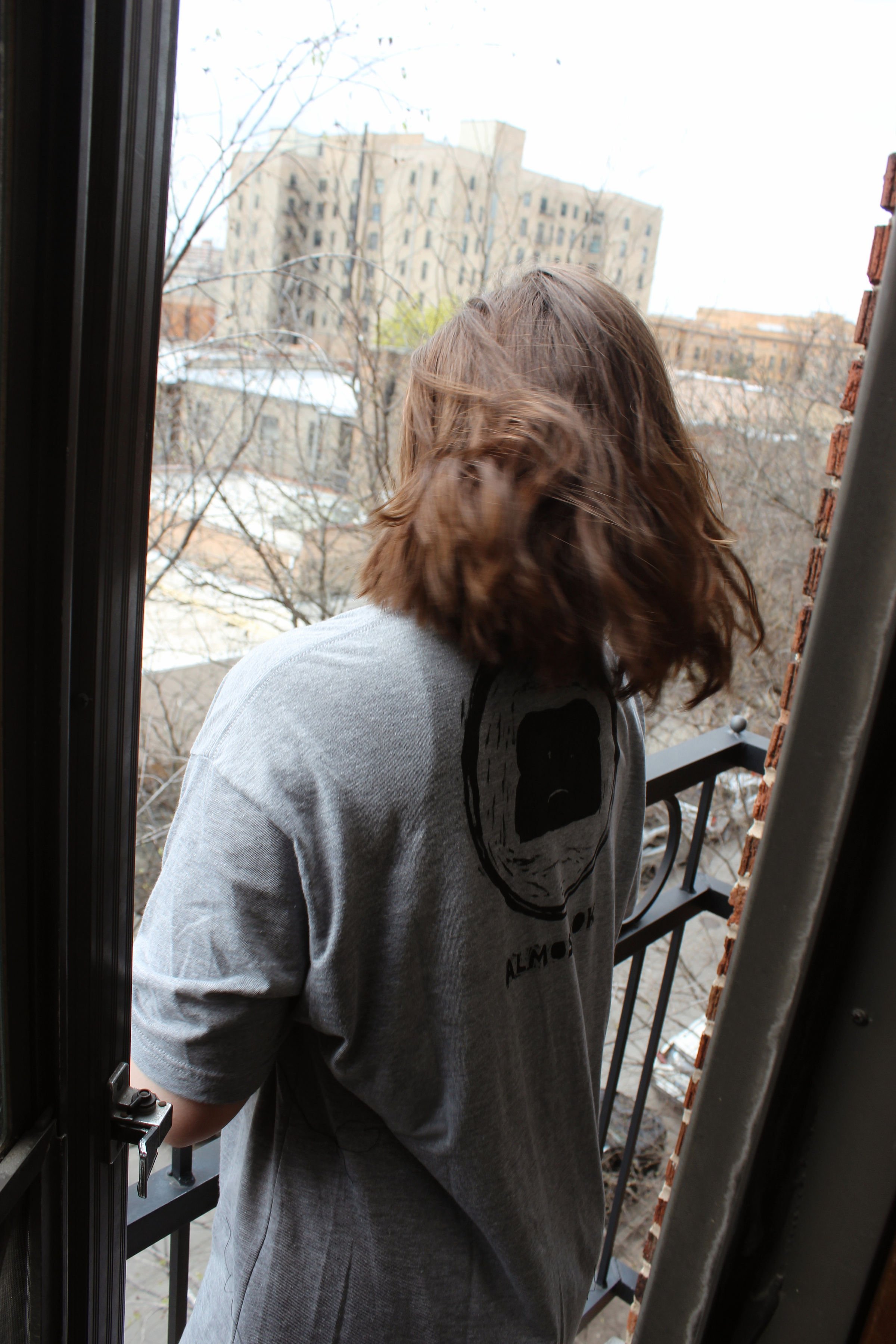 Person with wavy brown hair standing on a balcony, facing away, with a cityscape of tall buildings and leafless trees in the background. She is wearing a hand printed oversized T-shirt.