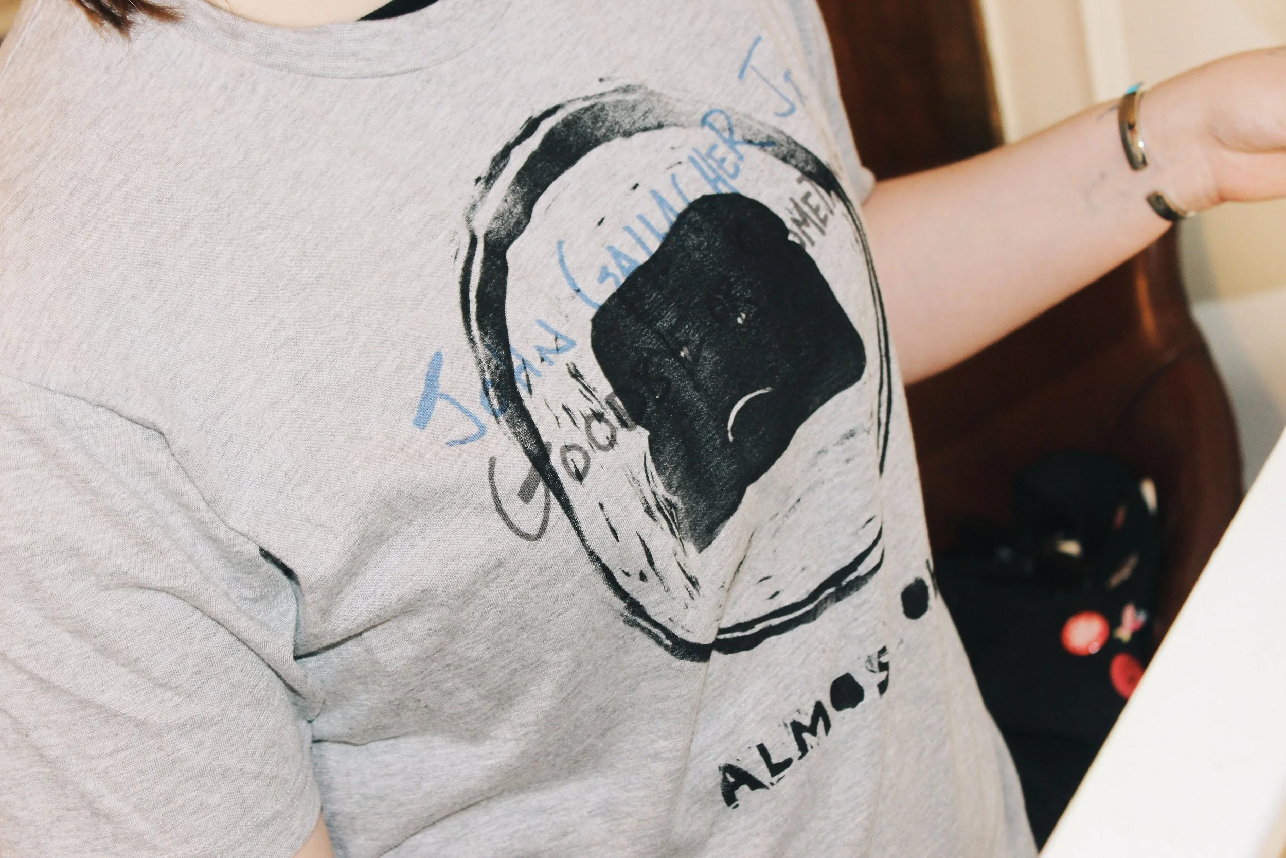 Person wearing a light gray T-shirt with a black handmade graphic and text around it, sitting at a table in an indoor setting.