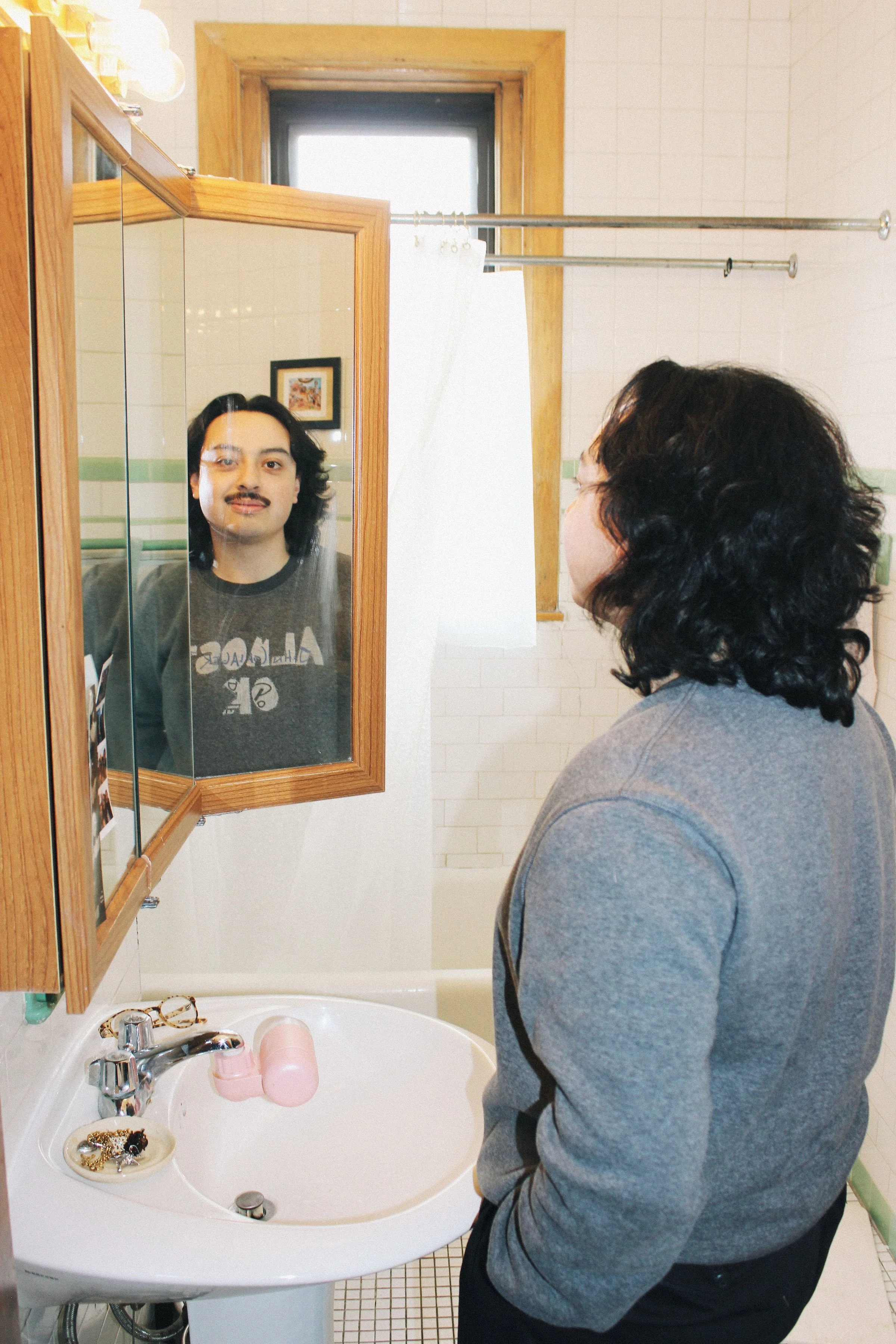 Person looking into a bathroom mirror with a mustache, wearing a gray sweatshirt with patches, in a bathroom with a small vanity and a window.