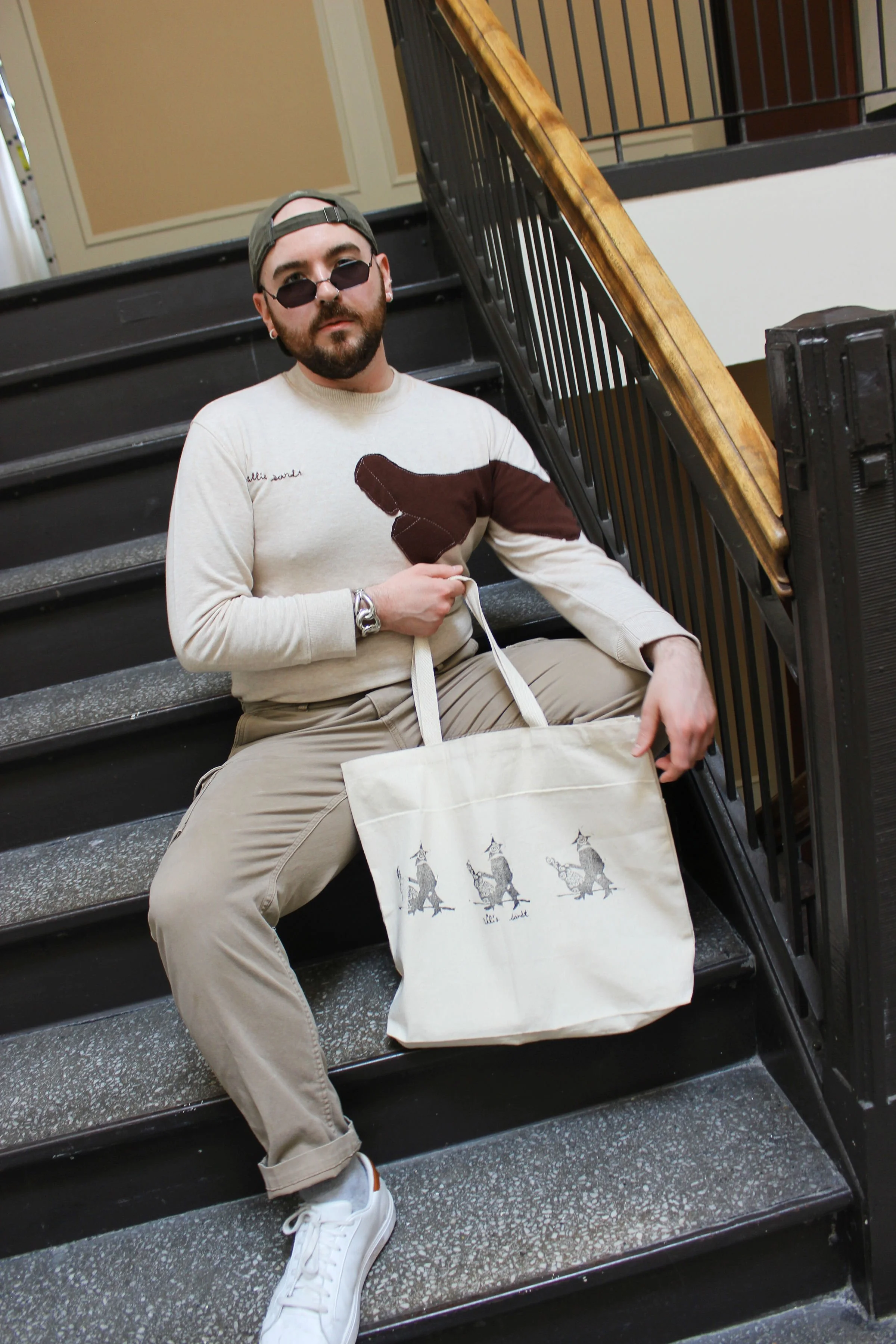 A man with a beard, sunglasses, and cap sitting on black stairs, holding a tote bag with three illustrated figures.