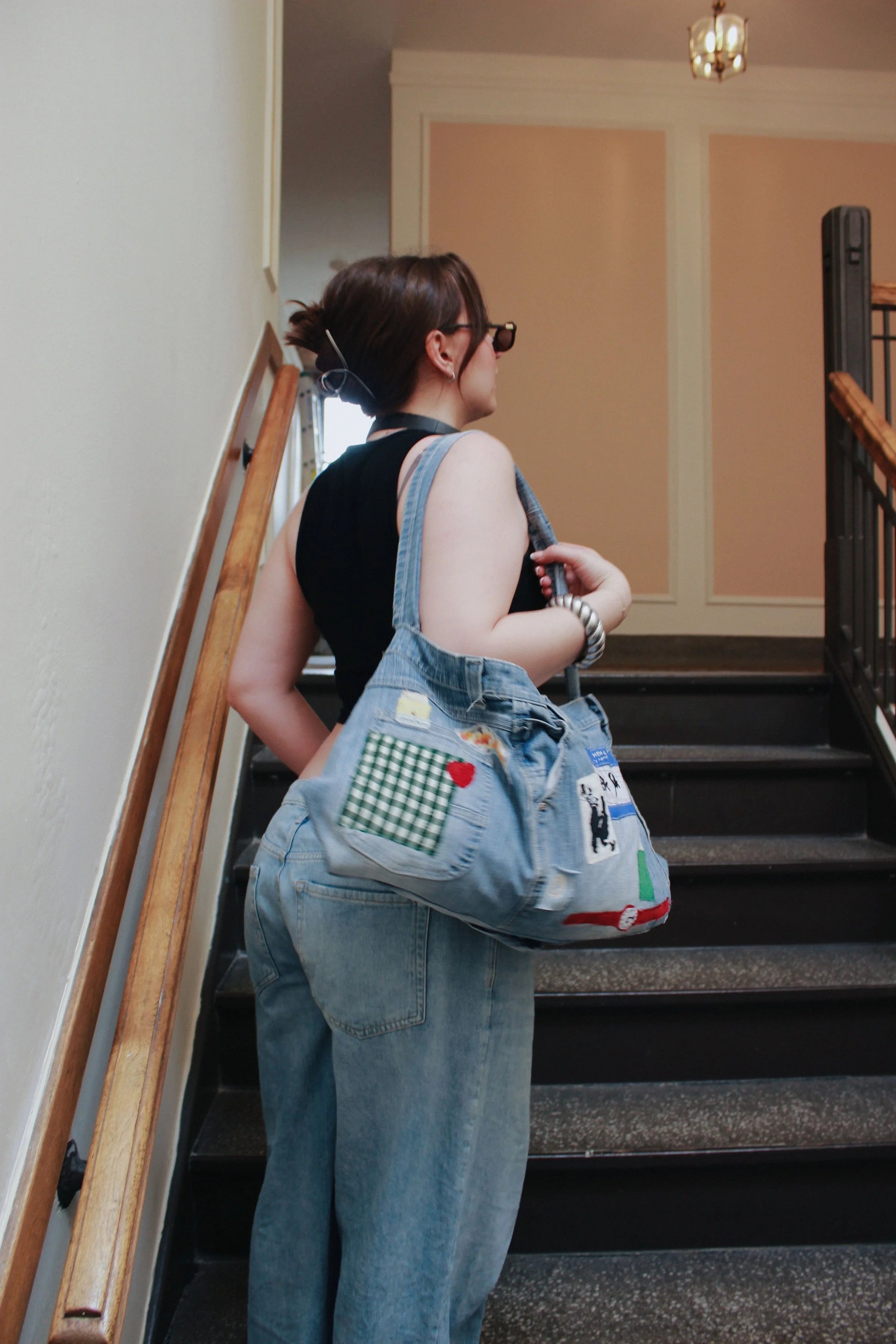 A woman with short hair and sunglasses carrying a denim bag decorated with patches, standing on a staircase and looking to the side.