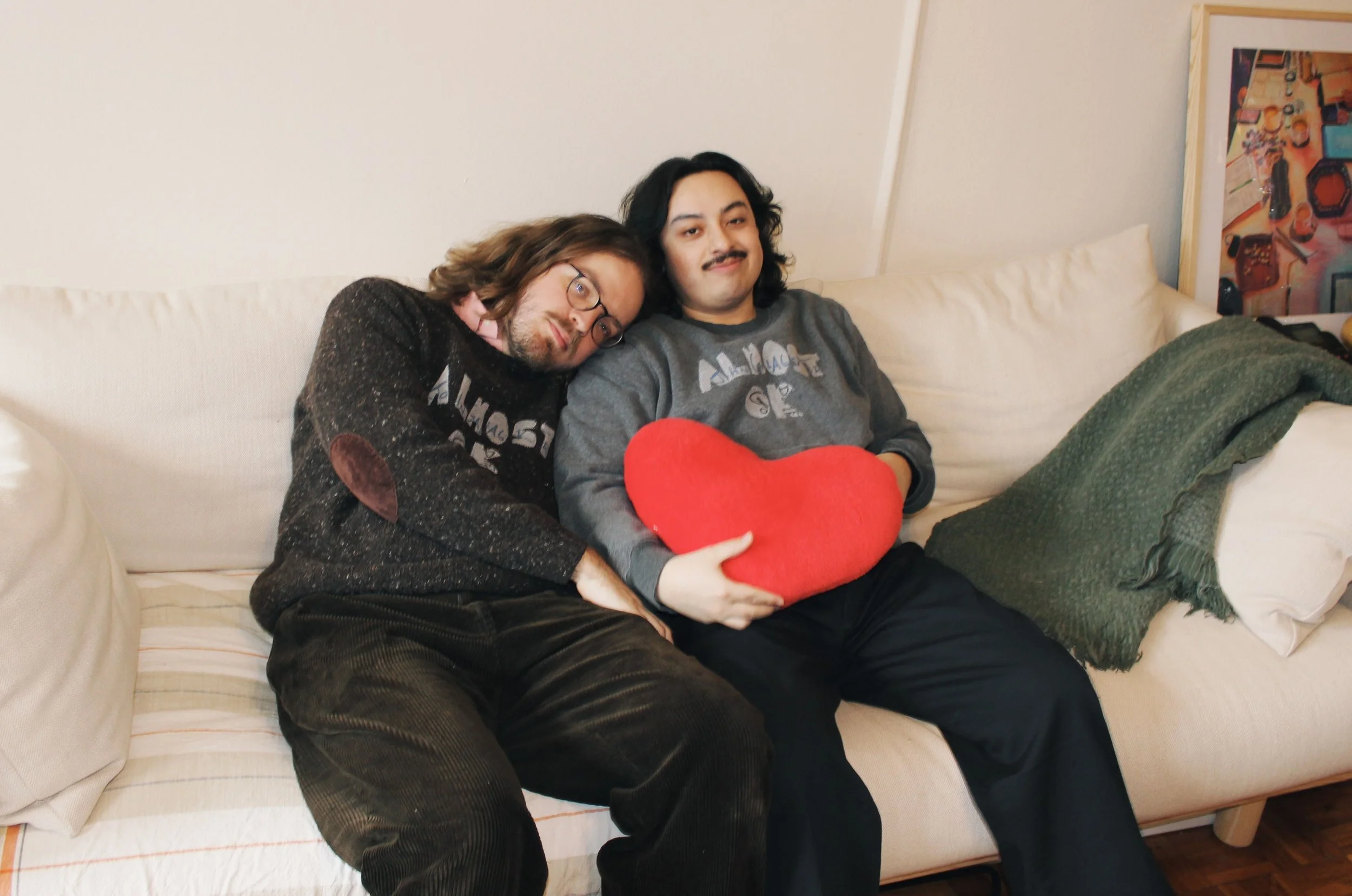 Two men sitting on a cream-colored sofa, one leaning his head on the other's shoulder. The man on the right is holding a red heart-shaped pillow, and they both appear relaxed and content.