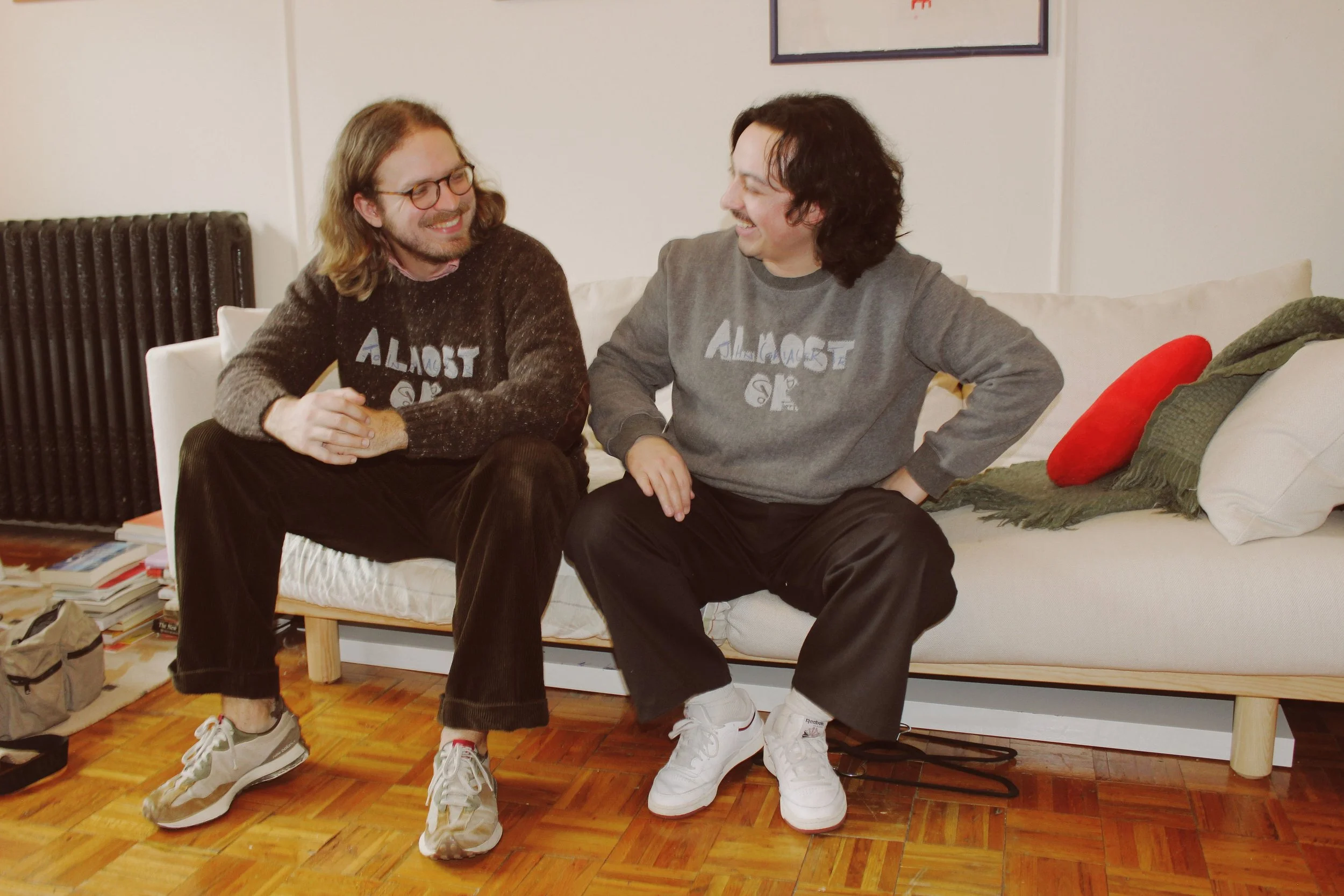 Two young men sitting on a white couch, smiling and engaging in a conversation. The man on the left has long hair, glasses, and is wearing a dark sweater, while the man on the right has curly hair and is wearing a gray sweatshirt. The room has hardwo
