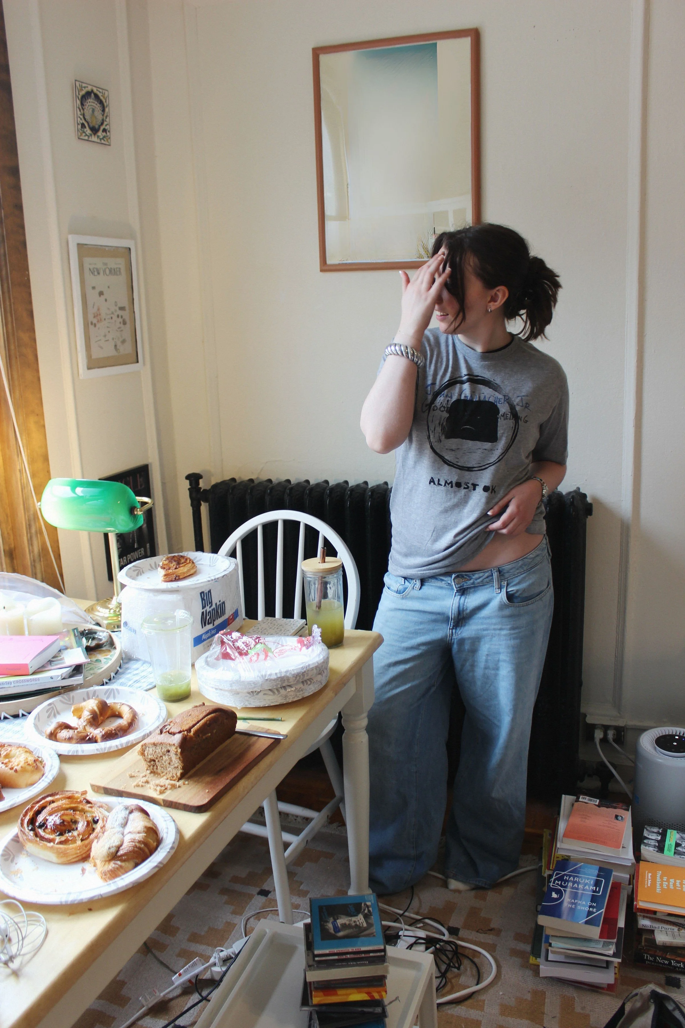 A young woman with dark hair tied back, wearing a gray T-shirt and loose jeans, standing in a cluttered room, touching her forehead with her right hand, with snacks, drinks, books, and a small speaker on a table.