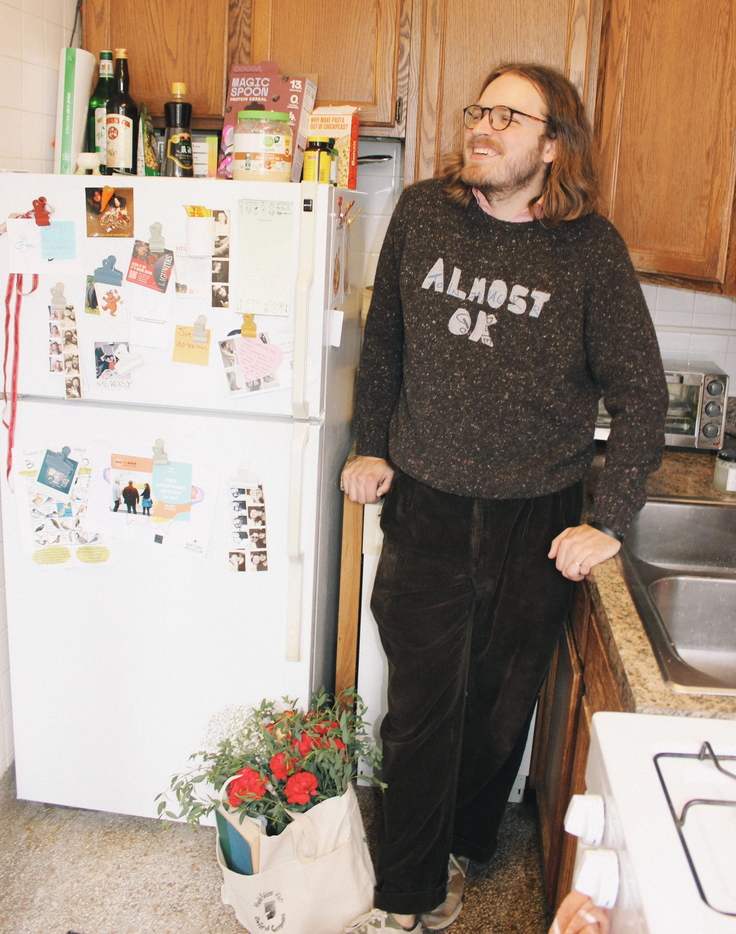 A man with glasses and long hair standing in a kitchen, smiling, with one hand on the counter and the other resting on a nearby cabinet. There is a white refrigerator with various pictures, notes, and magnets on it, and a pot with red flowers on the 
