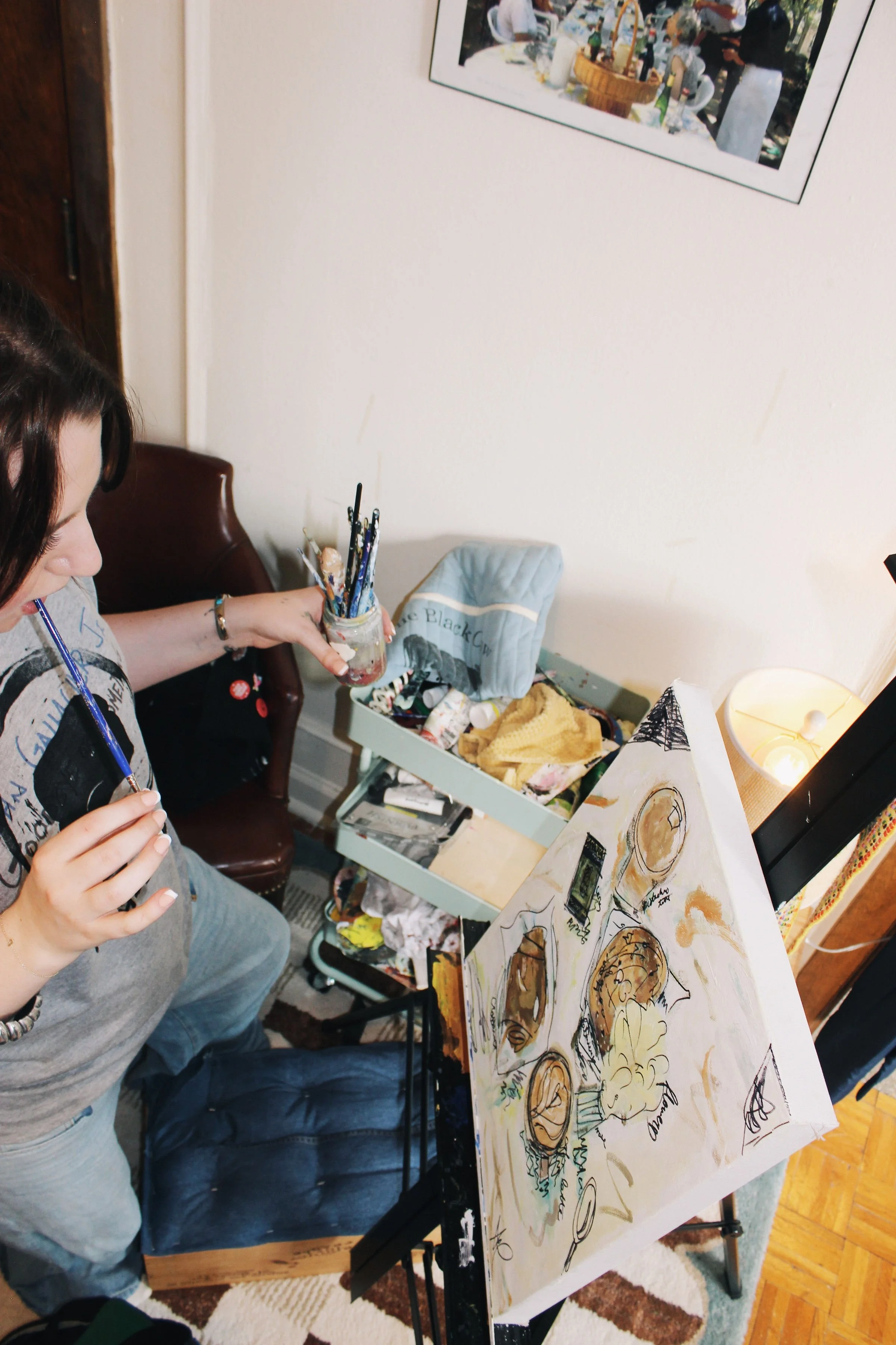 A person painting a food-themed artistic canvas on an easel in a cozy room, with art supplies and a cart nearby.