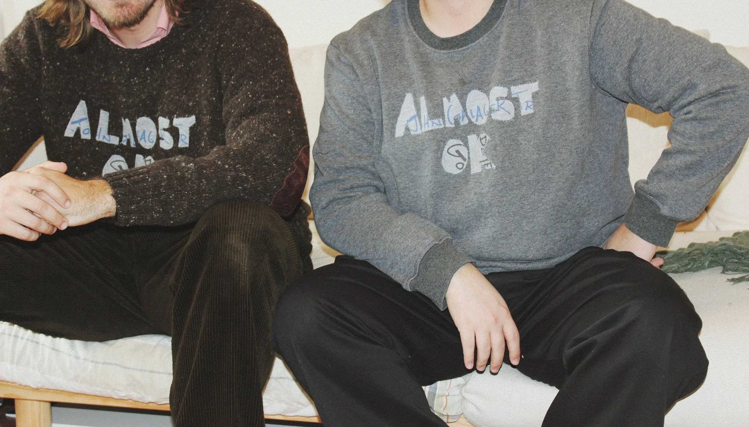 Two people sitting on a couch, wearing sweaters with the word "ALMOST OK" written across the front with patches.